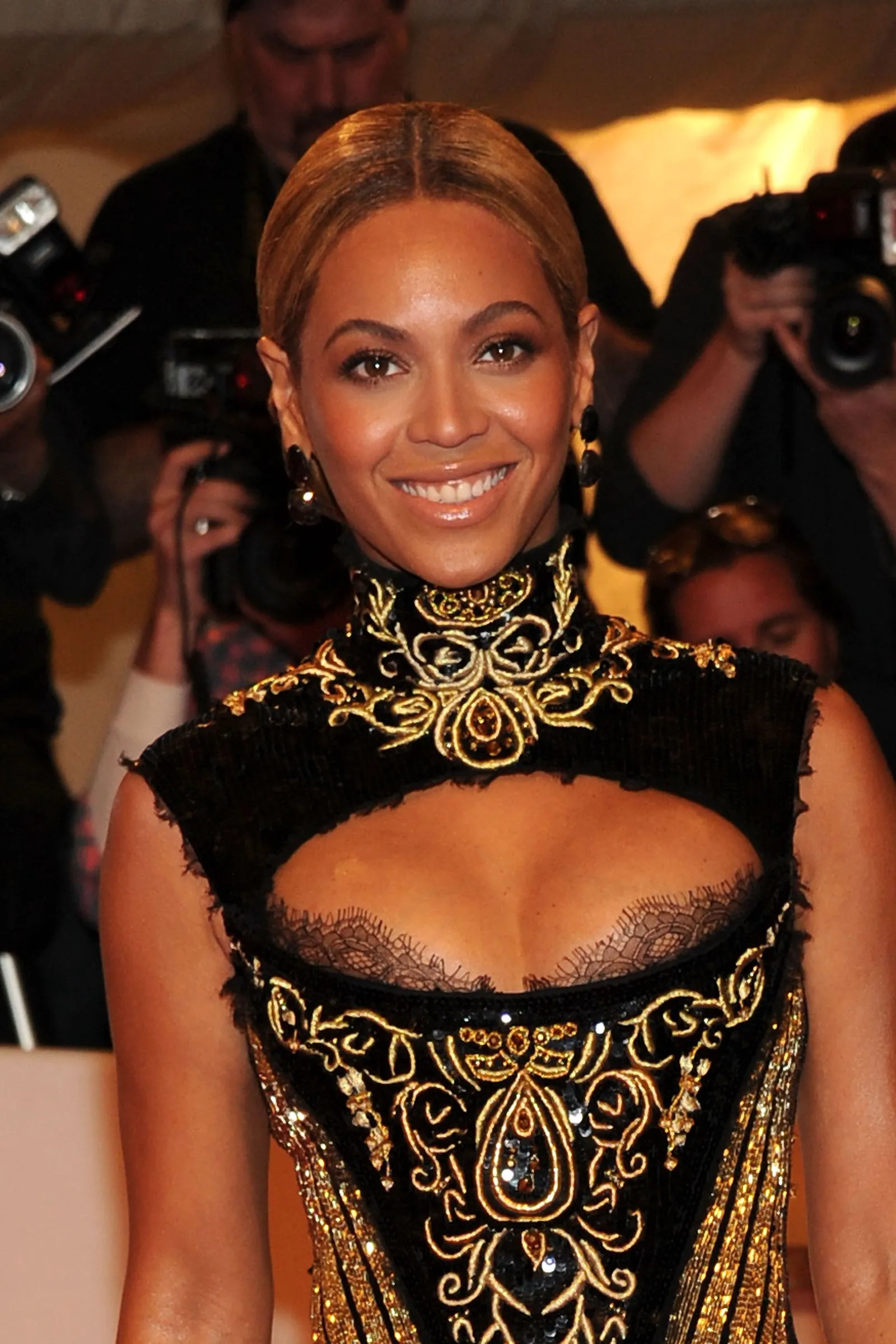 Smiling woman in a black and gold embroidered dress at a media event, photographers in the background.