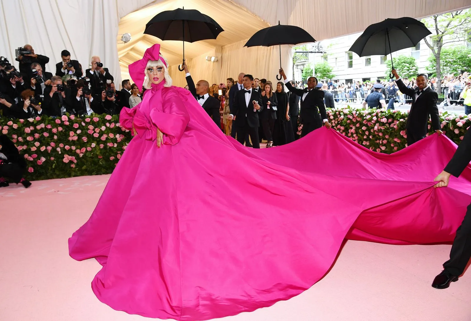 Person in a bright pink, voluminous gown with an enormous train on a pink carpet, surrounded by photographers and guests under a canopy.