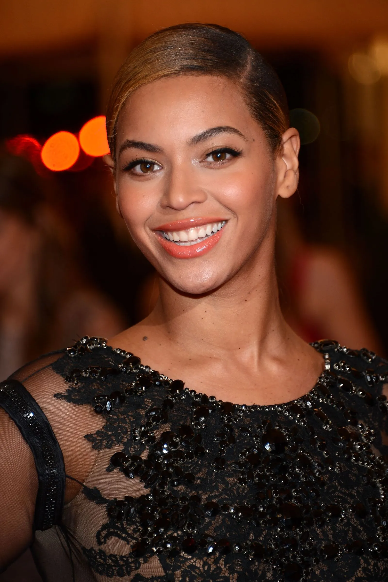 Smiling woman with slicked-back hair wearing a black lace dress with jeweled embellishments.