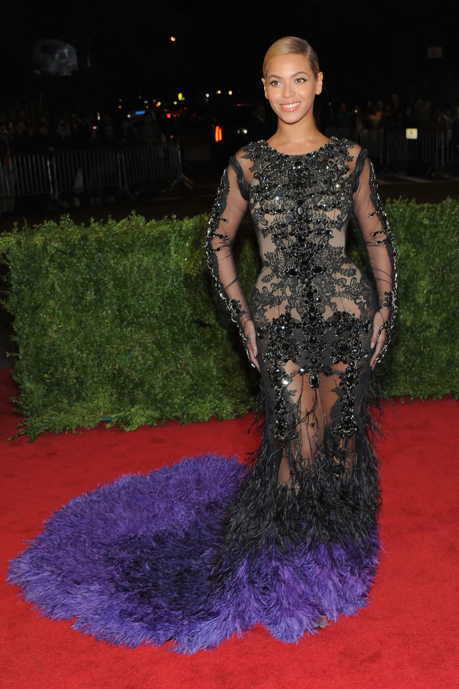 Woman posing on a red carpet in a black beaded lace evening gown with sheer panels at night.