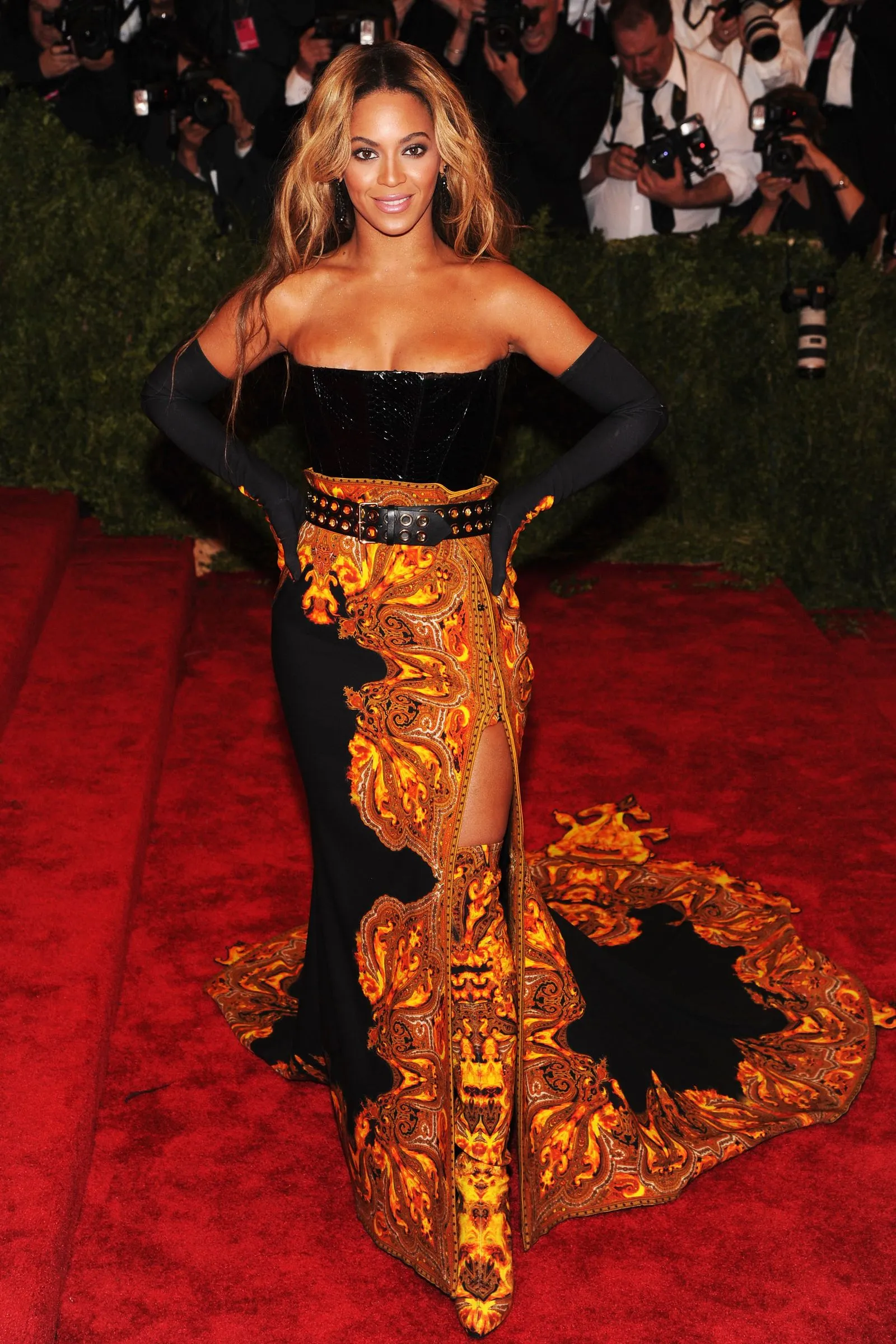 Woman posing on a red carpet in a black off-shoulder dress with a fiery orange-gold patterned skirt and studded belt, photographers behind her.