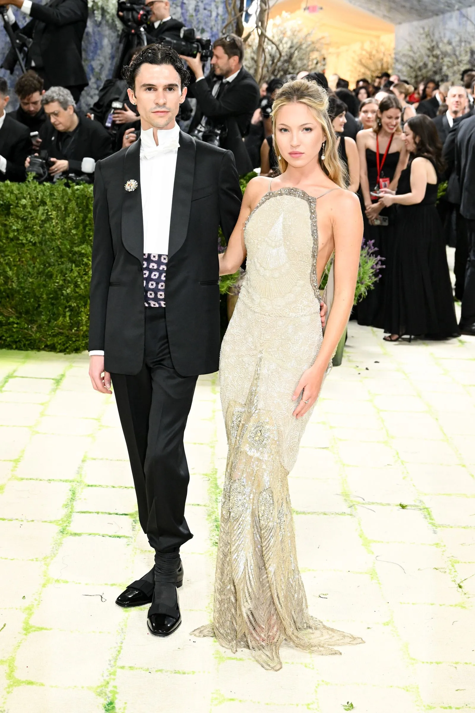 Man in a black tuxedo with a white bow tie walking arm-in-arm with a woman in a cream beaded gown at a formal event, photographers in the background.