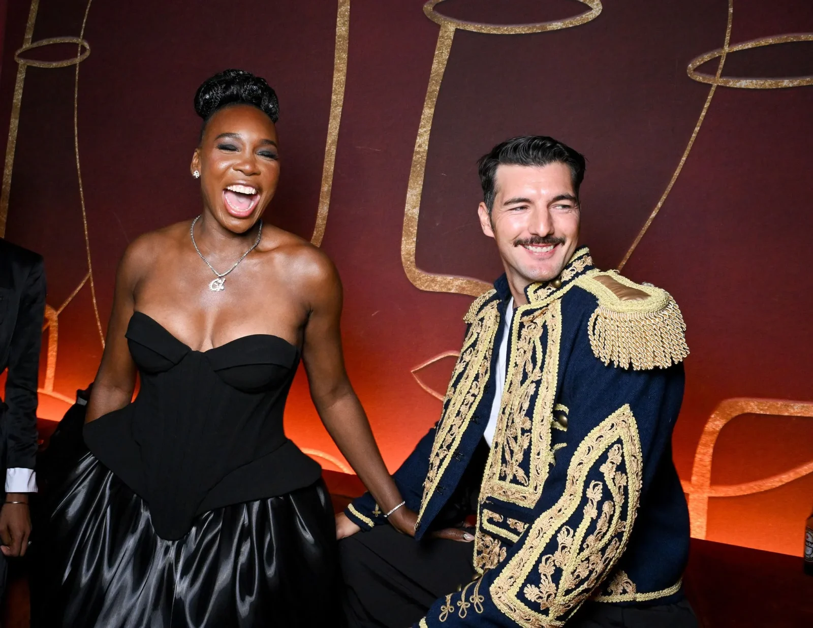 Two people posing and smiling against a red backdrop; woman in a black strapless dress, man in a navy jacket with gold embellishments.