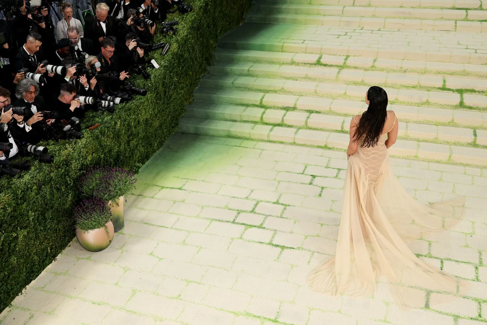 Crowd of photographers with cameras line the hedge wall, focusing on a woman in a long beige gown as she walks up stone steps at a formal event.