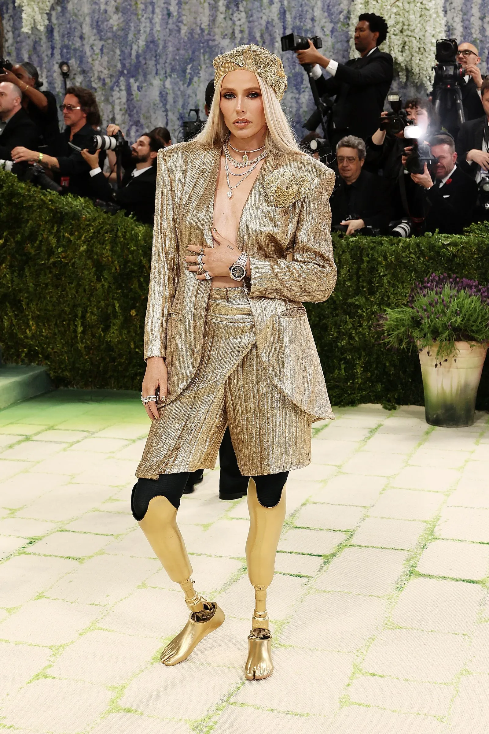 Woman in a shimmering gold suit and headpiece posing on a green carpet amid press photographers.