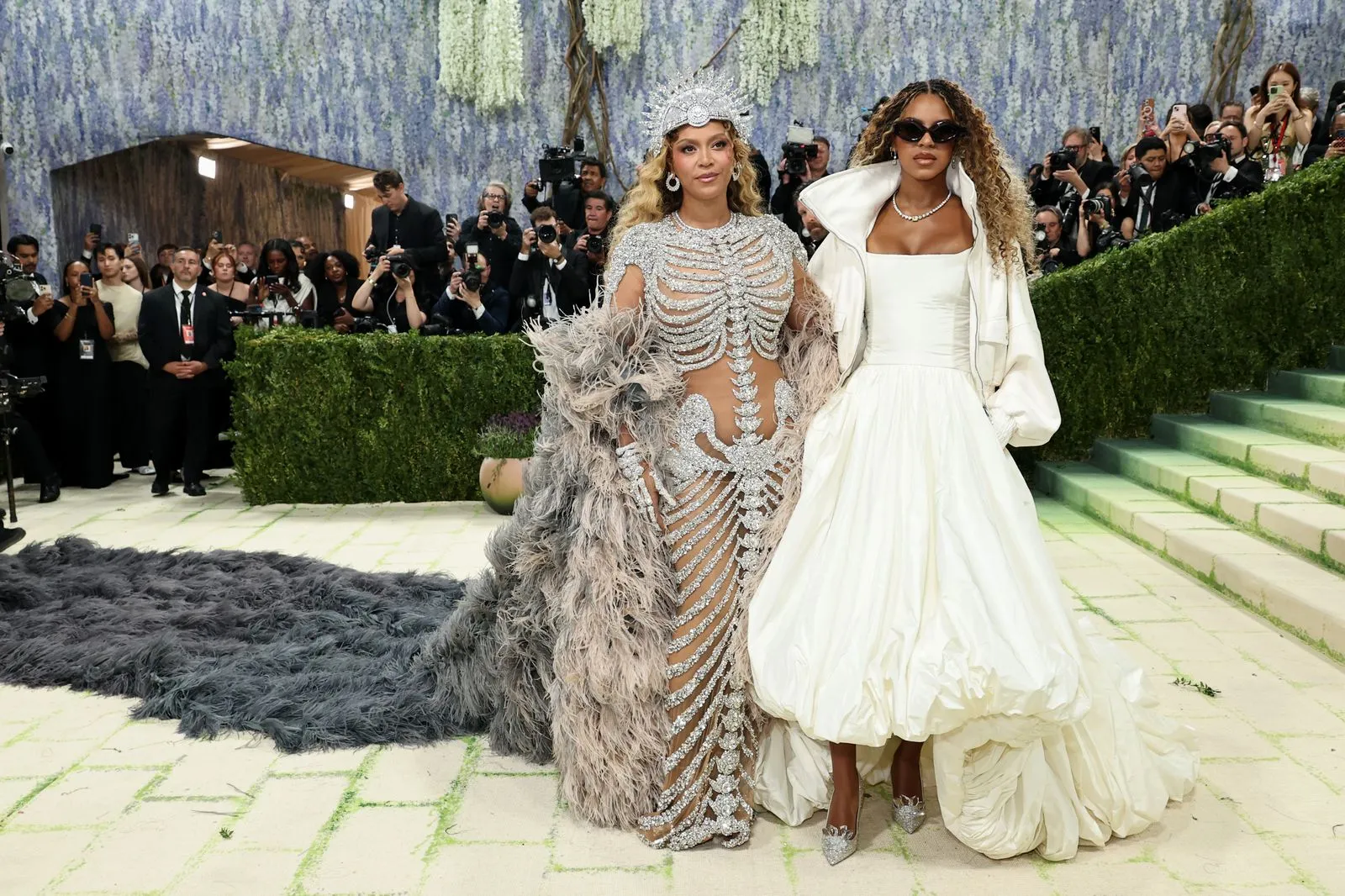 Two models pose on a red carpet: one in a silver, crystal-covered gown with a feathered cape; the other in a white off-shoulder gown with a large cape, as photographers line the background.