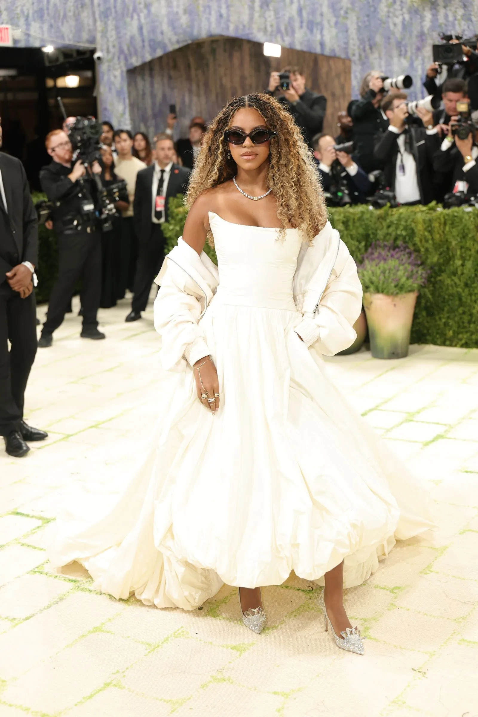 Woman in a white off-shoulder gown walking a red carpet with photographers in the background.
