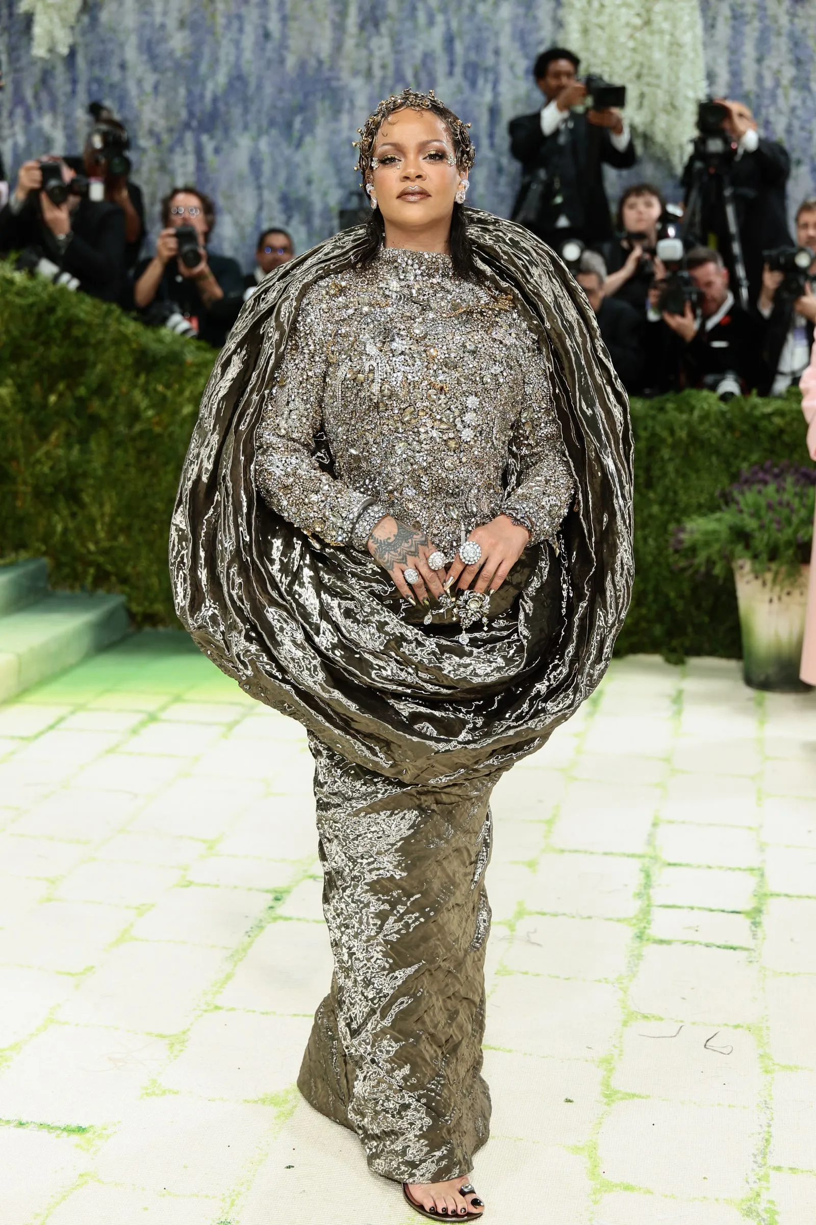 Woman wearing a heavily embellished silver gown with an oversized cape, walking on a fashion runway as photographers film in the background.