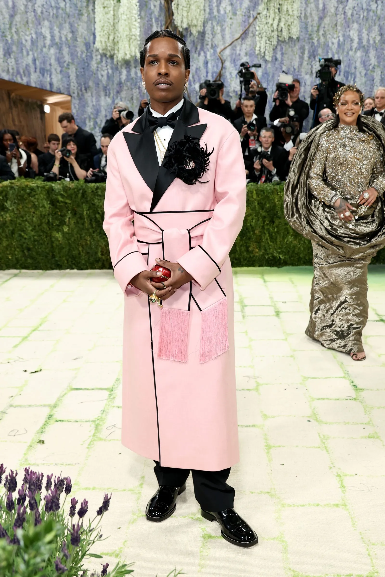 Man in a pale pink suit with black lapels and bow tie posing on a gala red carpet as photographers loom in the background, lavender floral backdrop behind