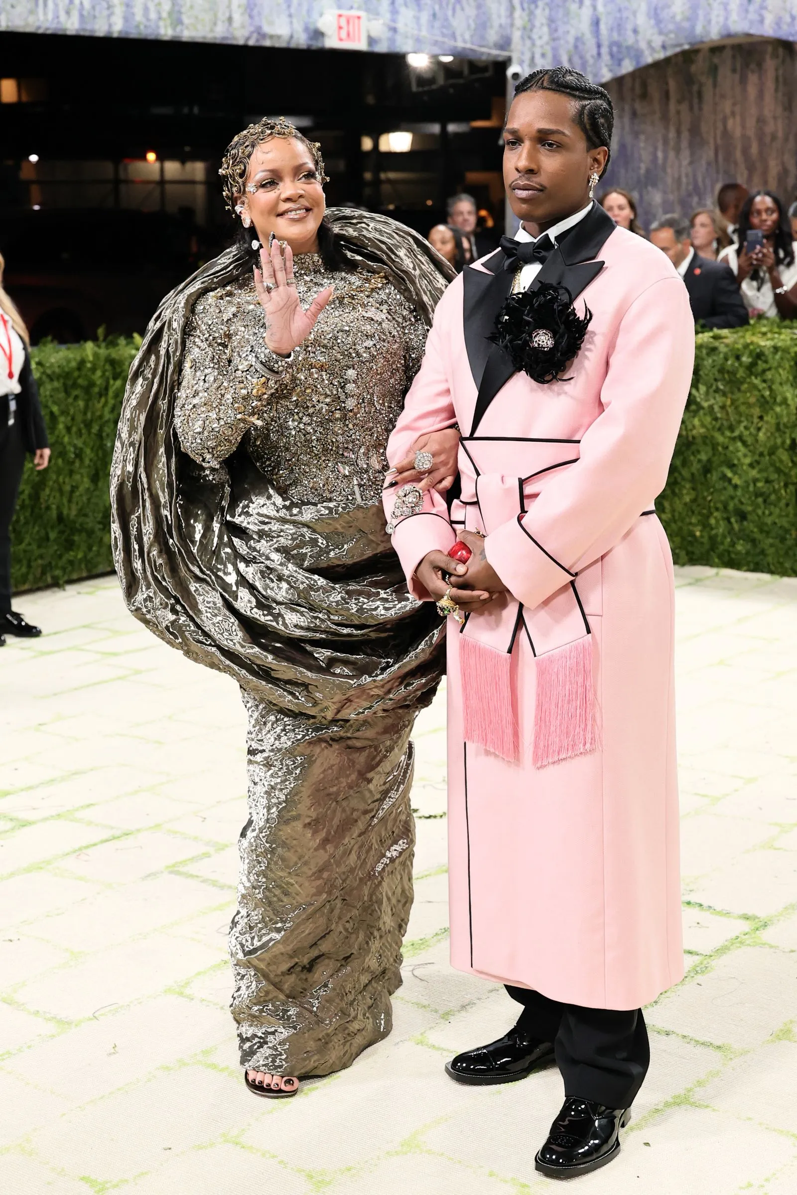 Woman waves beside a man in a pink tuxedo at a formal event, both dressed in elaborate, sparkling outfits.