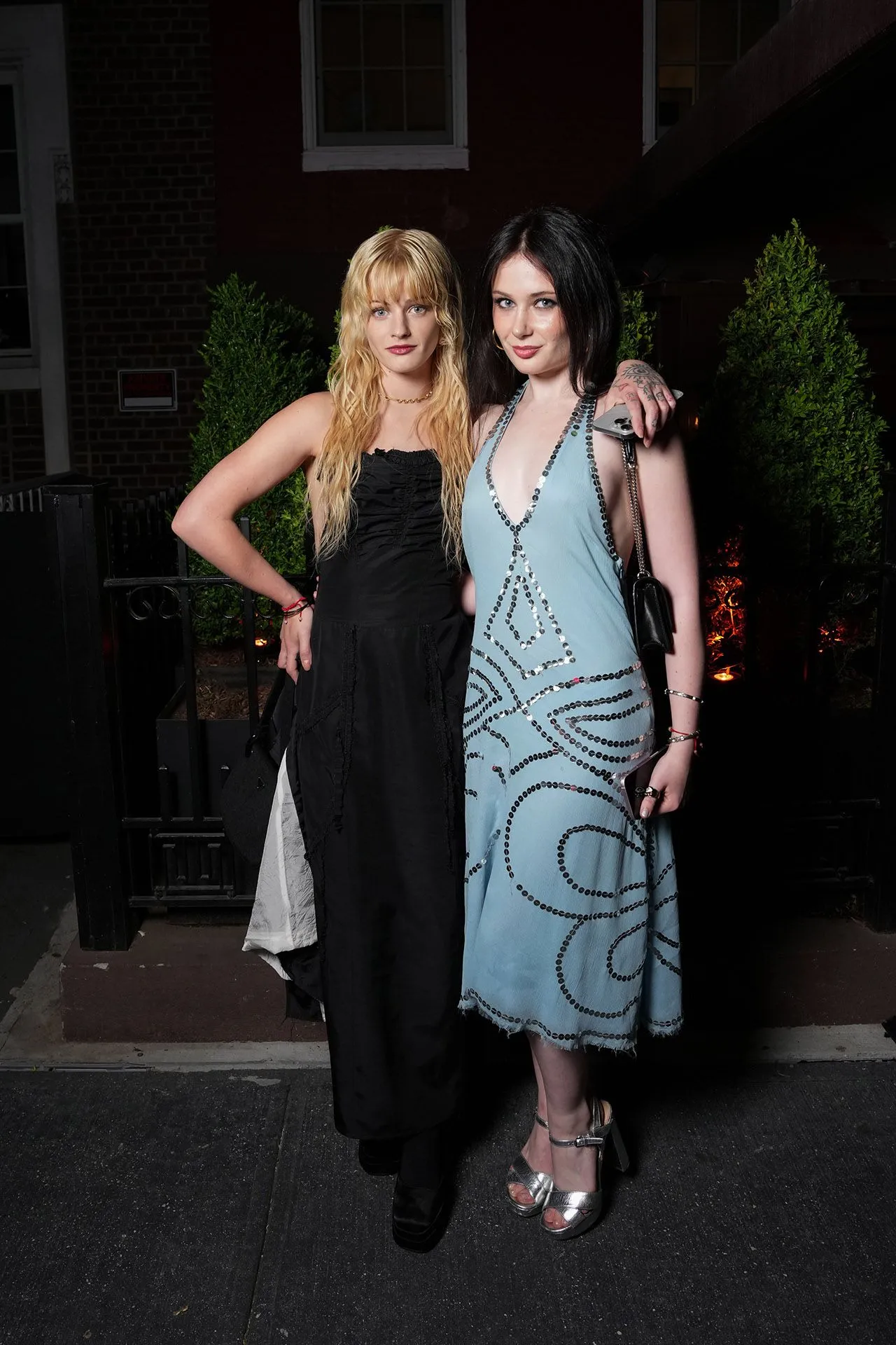 Two women posing together outdoors at night, one in a black dress and the other in a light blue beaded dress, arm around each other.