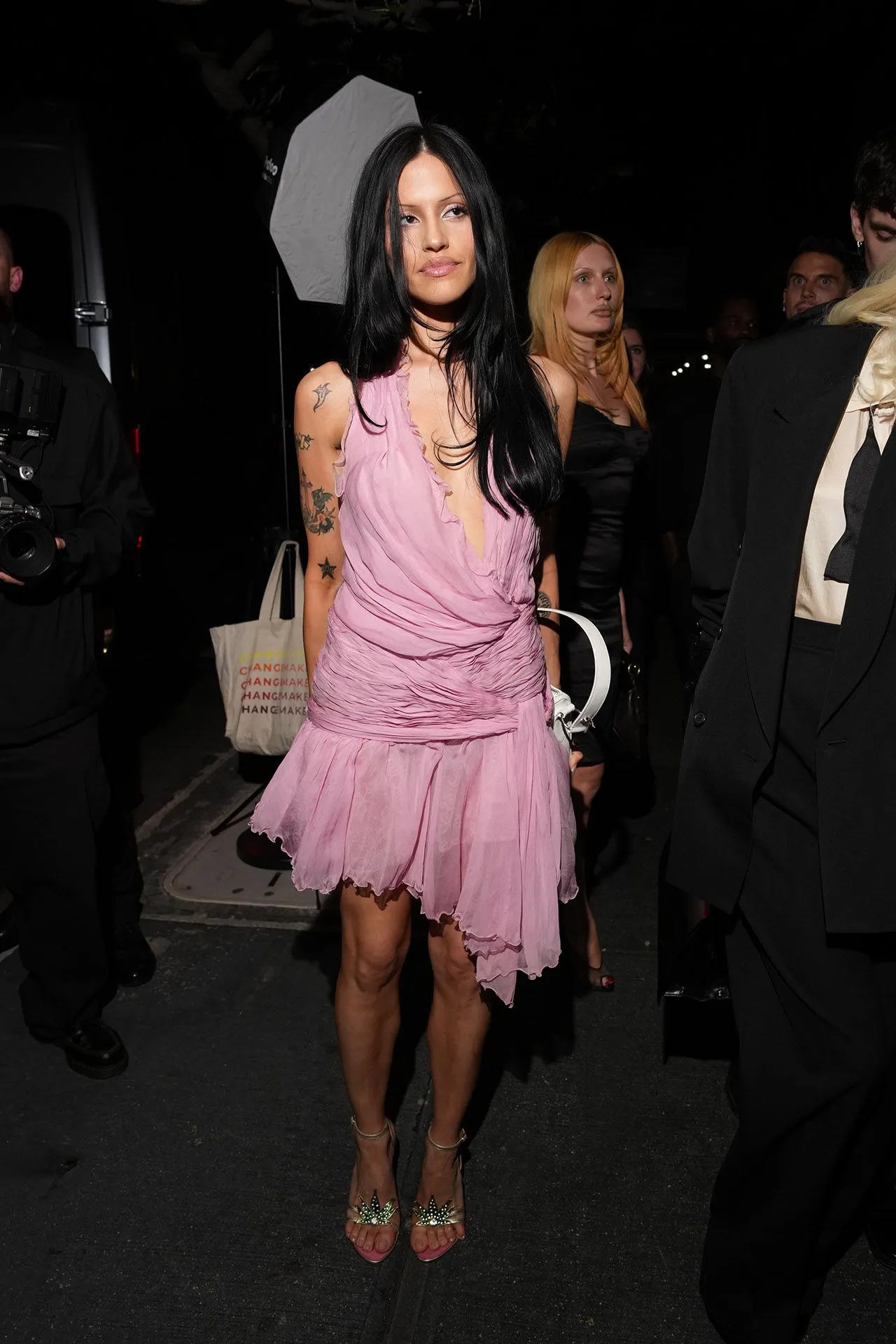 Woman with long black hair wearing a pink ruffled dress at night, surrounded by photographers and onlookers.