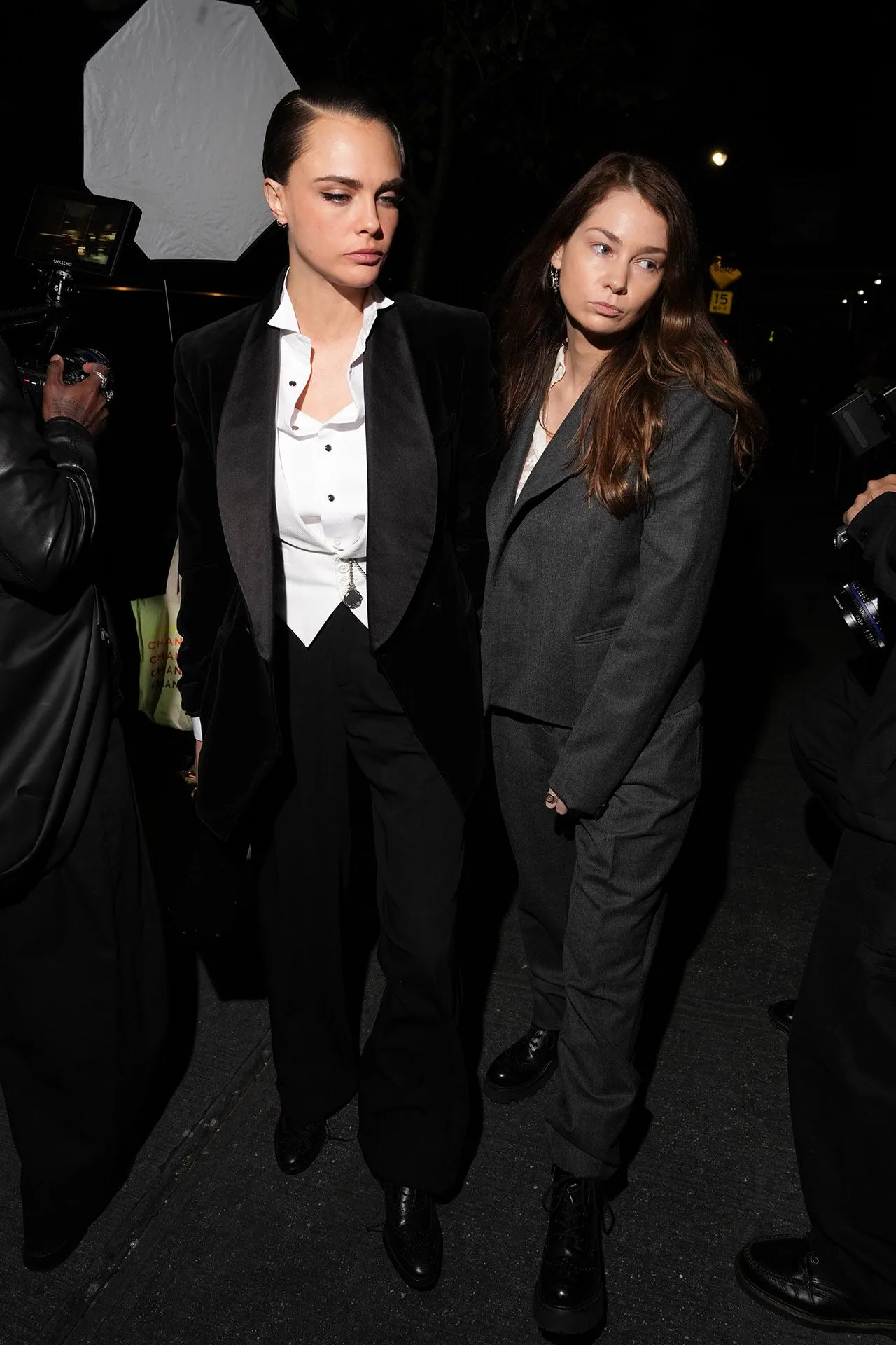 Two stylish women in dark formal outfits walk at night, surrounded by photographers and cameras on a city street at night.