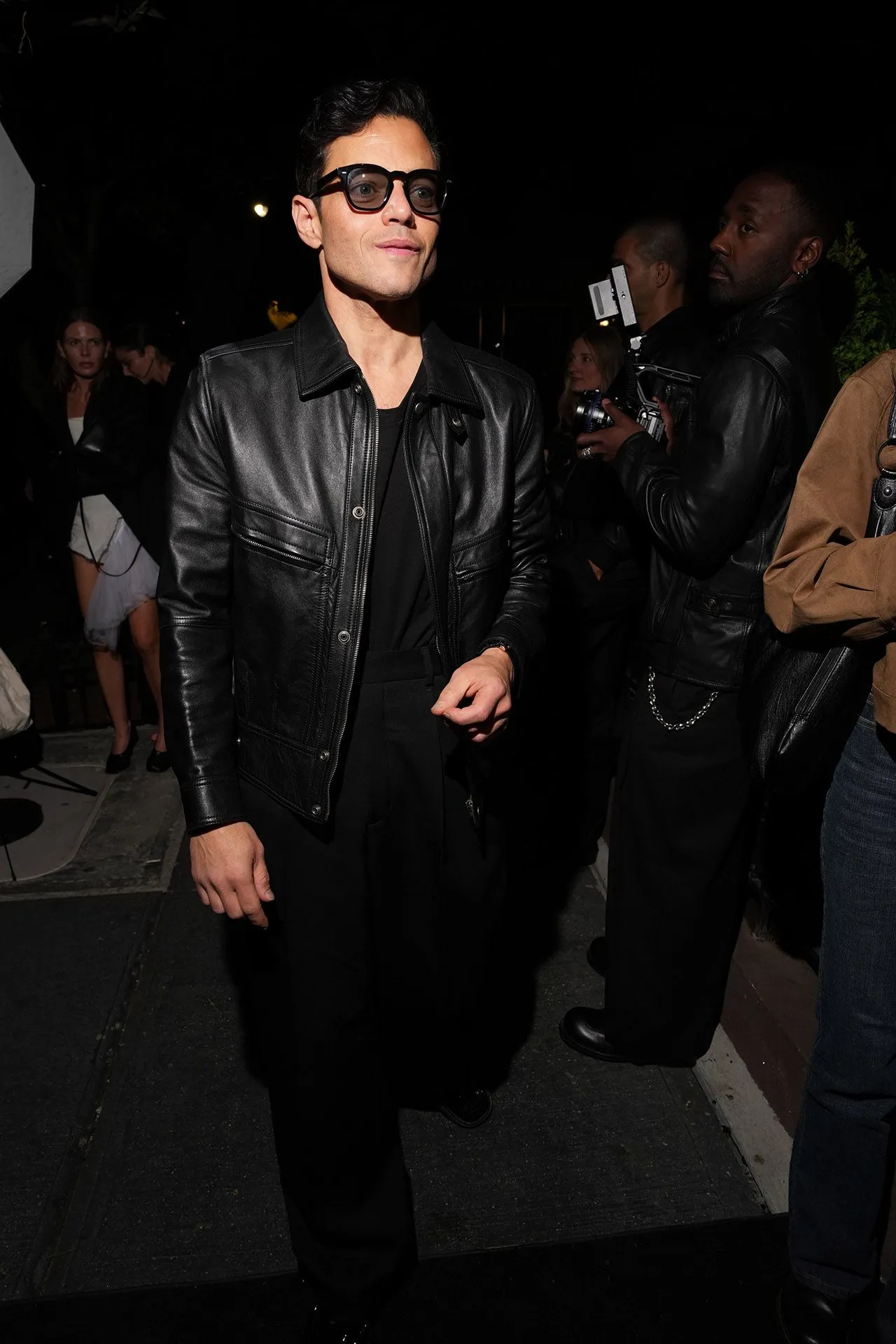 Man in a black leather jacket and dark glasses at a nighttime event, with photographers and other attendees in the background.