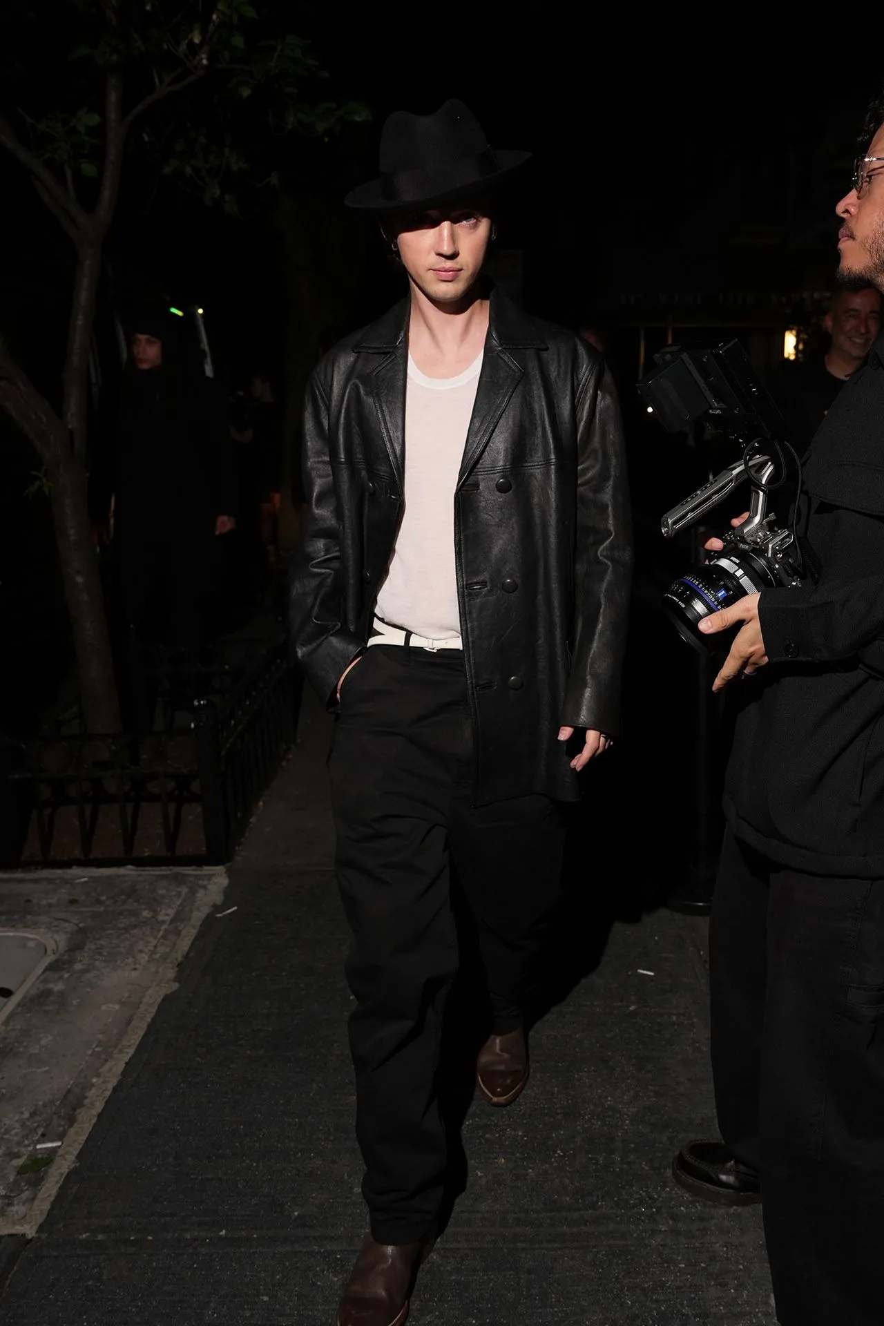 Man in a black leather jacket and wide-brim hat walks on a dim night street, a photographer with a camera beside him.