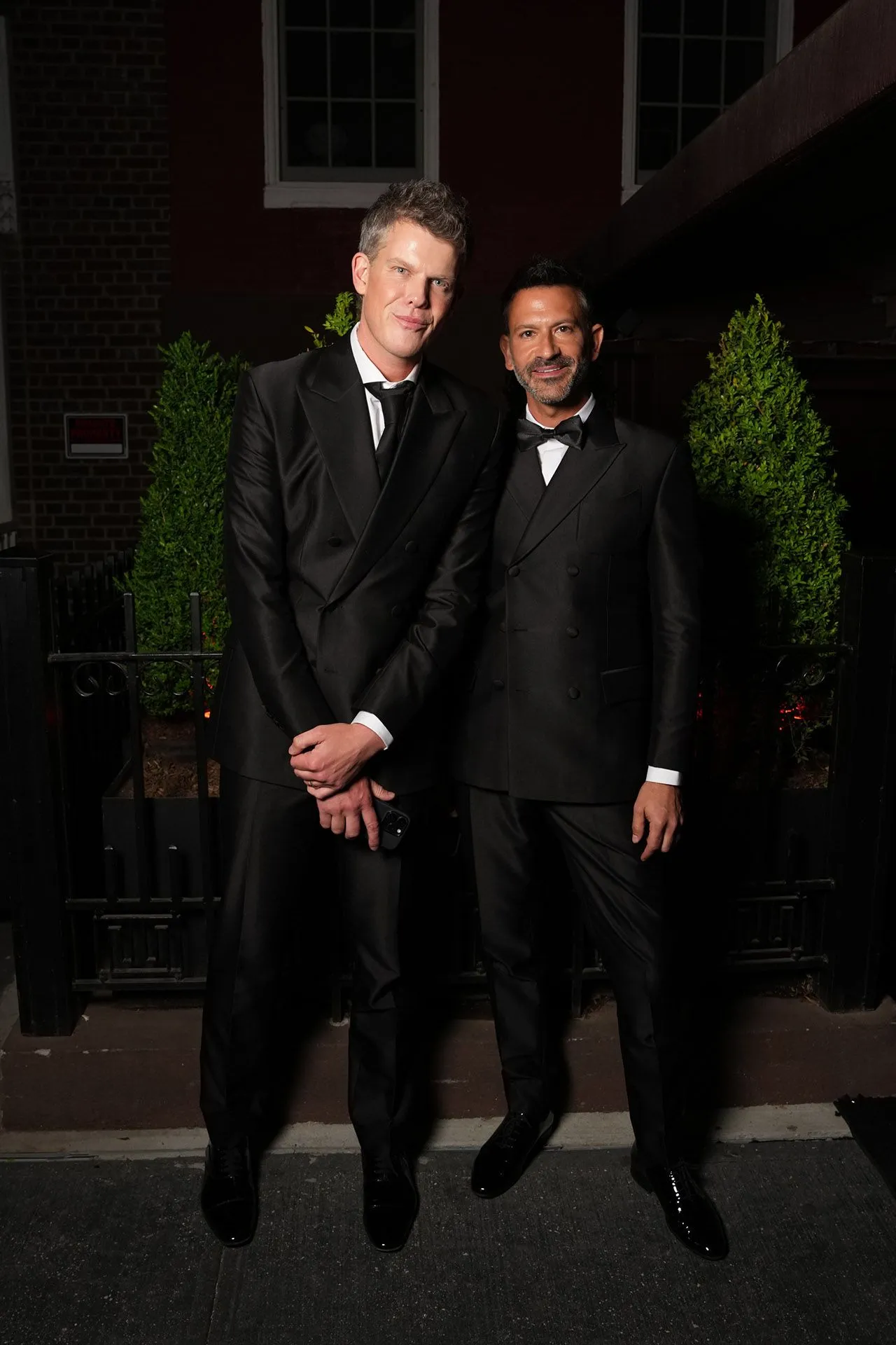 Two men in black tuxedos pose together outdoors at night beside bushes outside a building.