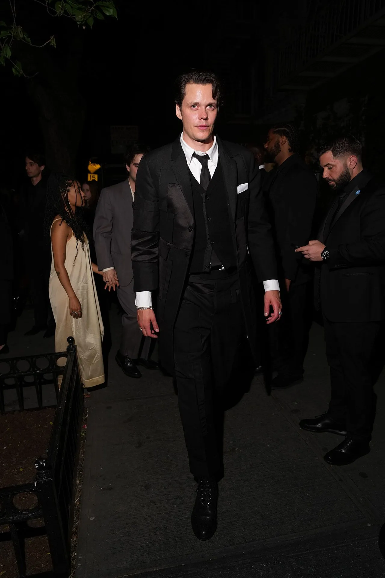 Man in a black suit and tie walking toward camera at night, others in evening attire in the background at a street event.