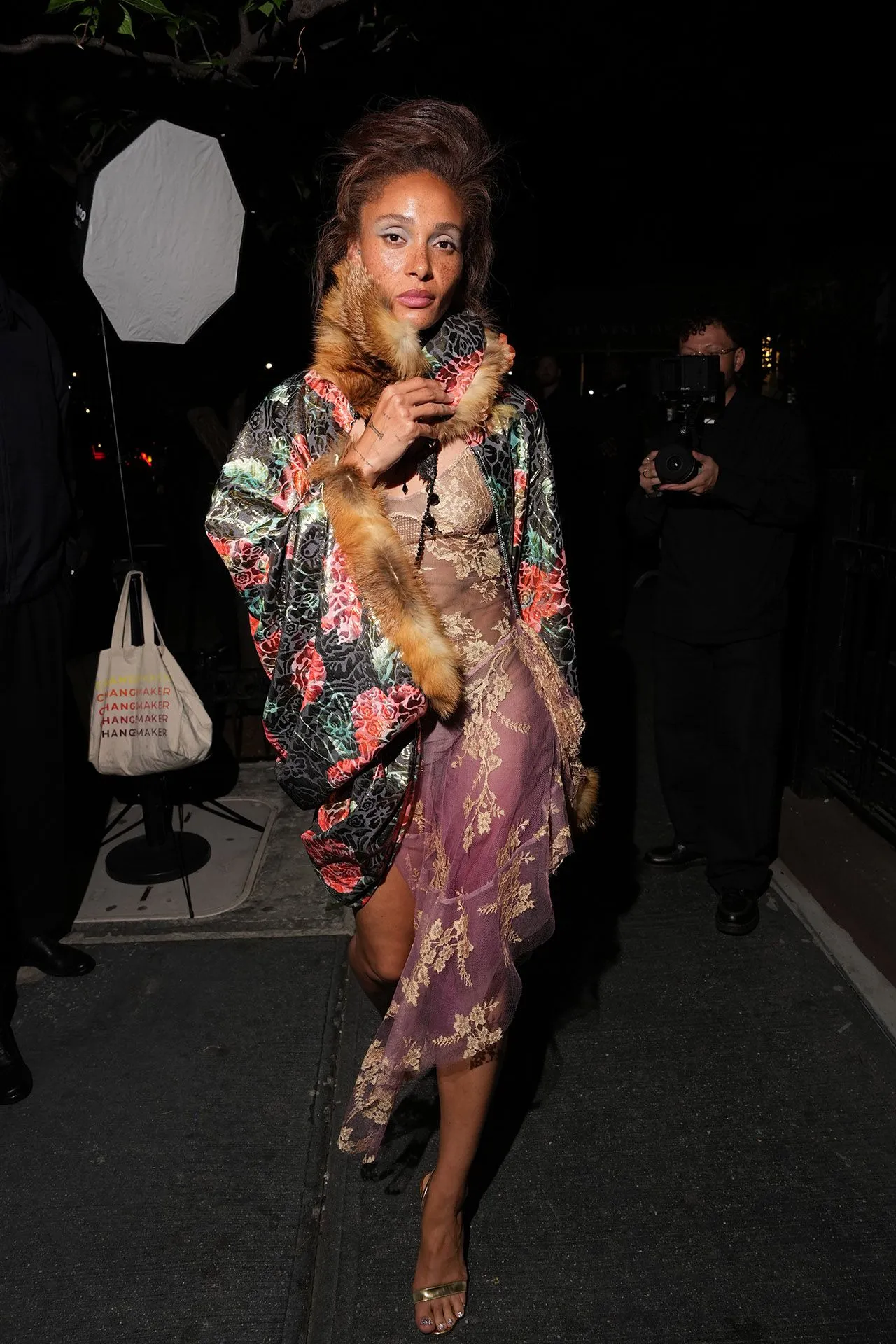 Model in a vibrant floral kimono with fur trim and a lavender lace dress, posing on a nighttime fashion set with a photographer in the background.