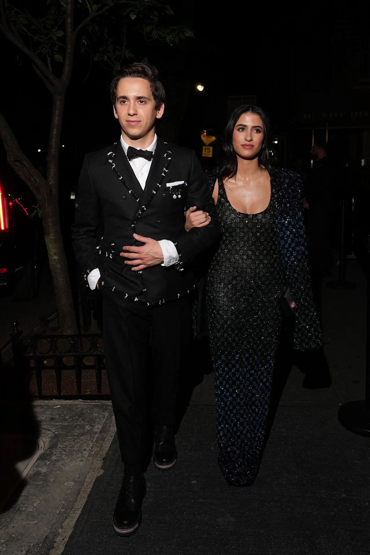 Two formally dressed individuals walking arm in arm at night; man in a black suit with a bow tie, woman in a glittery sequined outfit.