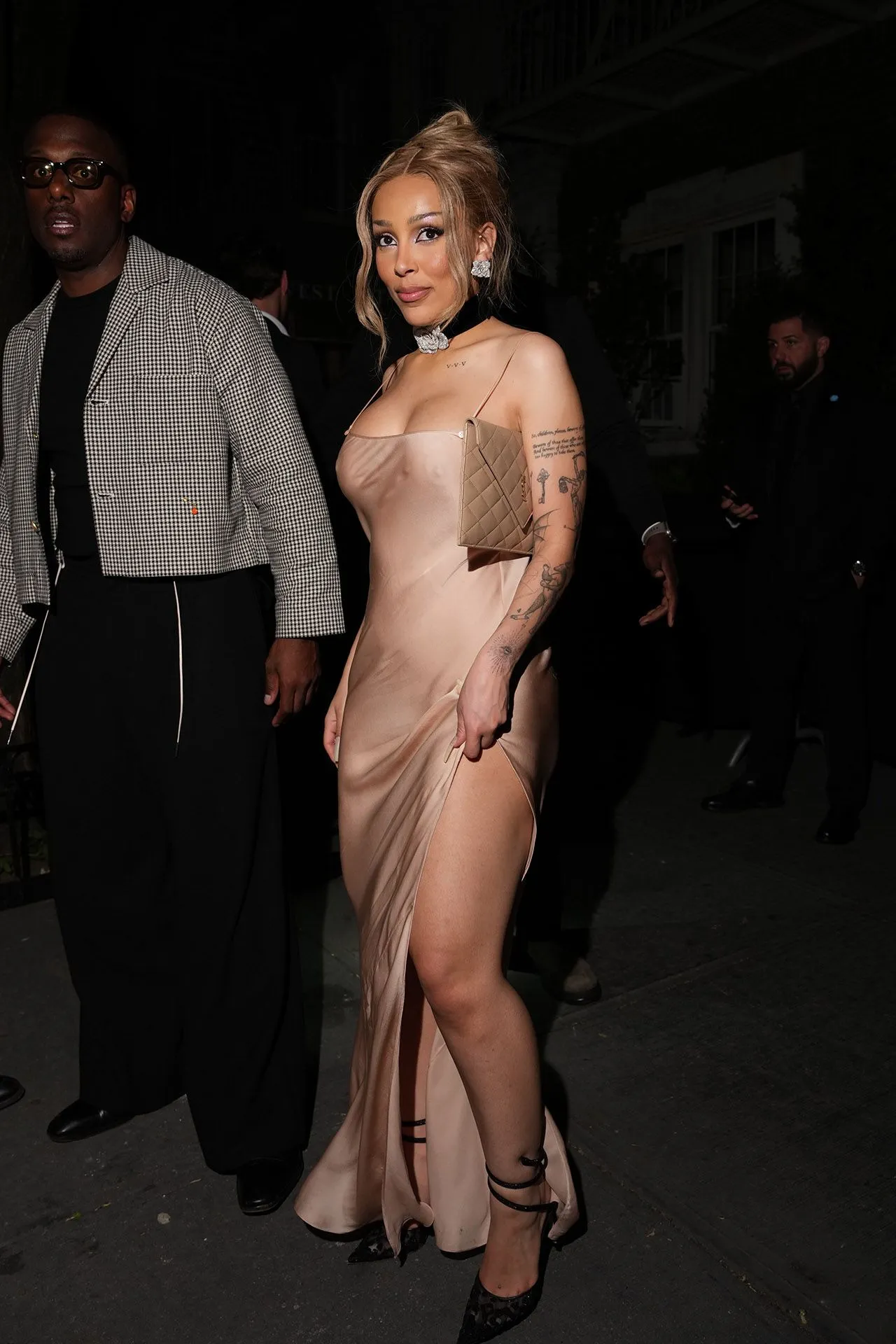 Woman in a silky champagne gown with a thigh-high slit, clutch under arm, walking at night, tattoos visible on her arm.