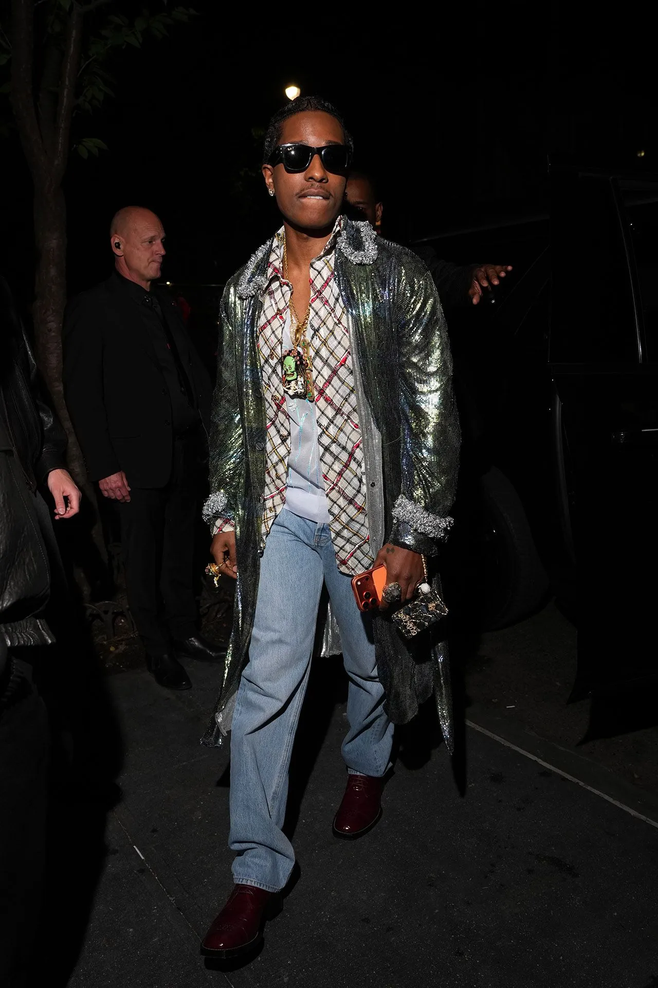 Man wearing a shiny metallic coat, plaid shirt, jeans, sunglasses, and multiple necklaces at night, holding an orange phone.