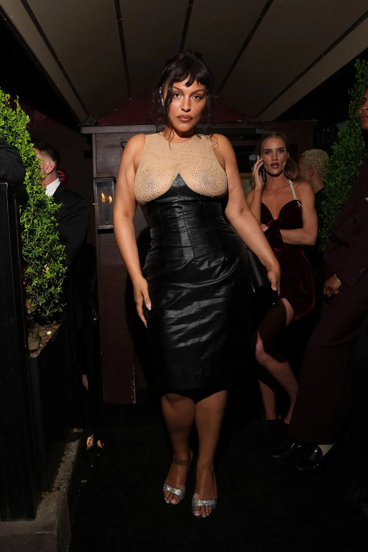 Woman in a beige rhinestone-embellished bodice and black satin dress walks down a dimly lit path at night, others in formalwear nearby.