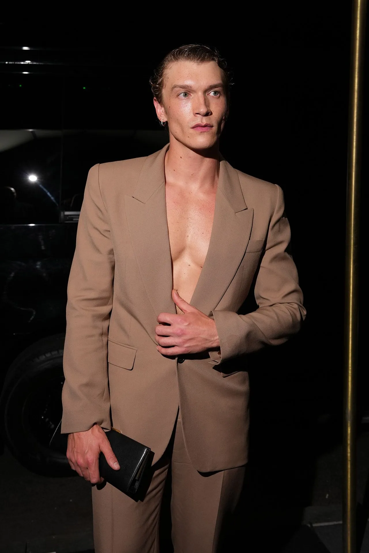 Man in a tan suit with a plunging jacket opening, holding a black clutch at his side, posing against a dark background at night