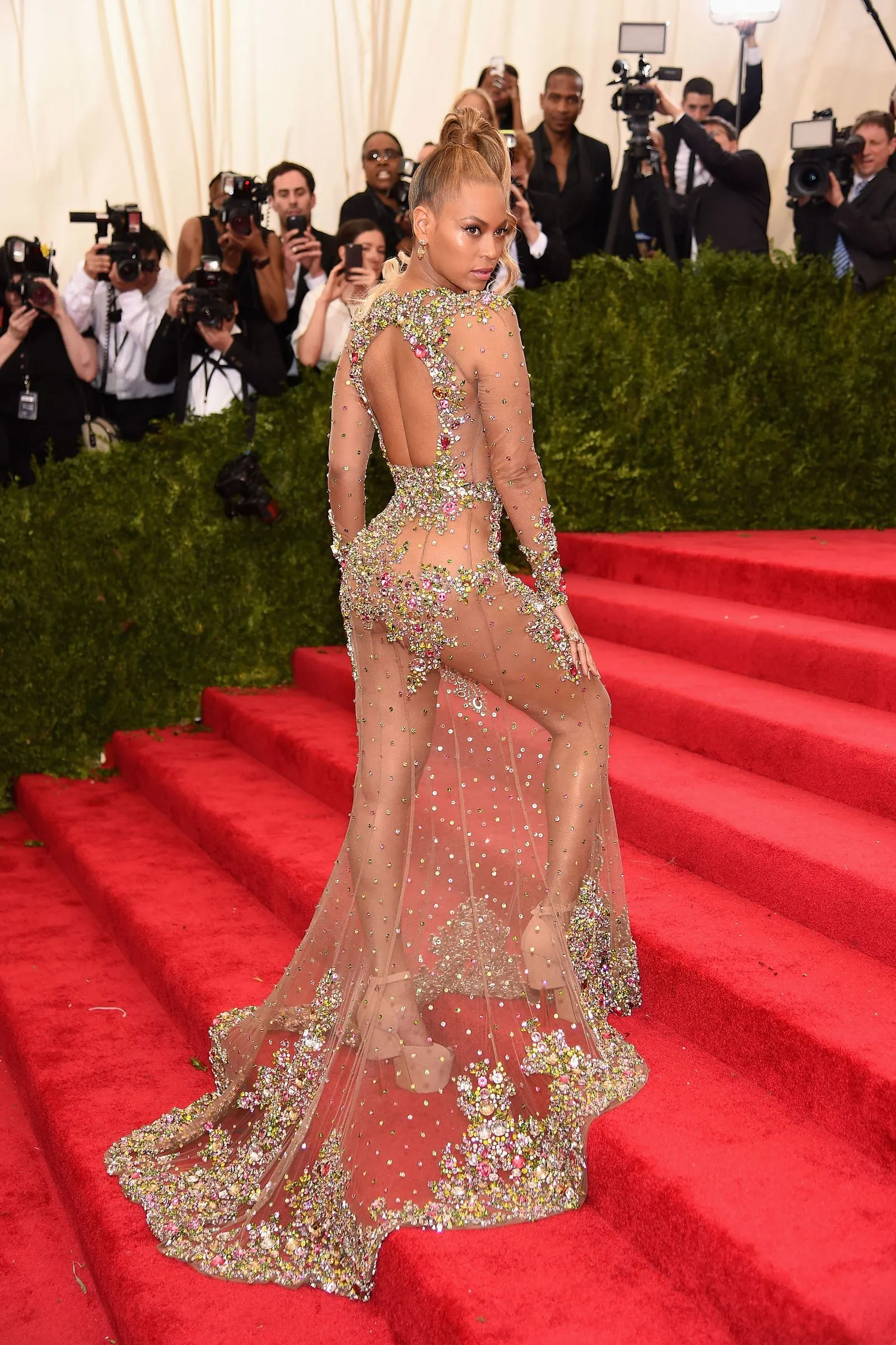 Celebrities posing on a red carpet; woman in a sheer, crystal-embellished gown with an open back, surrounded by photographers.