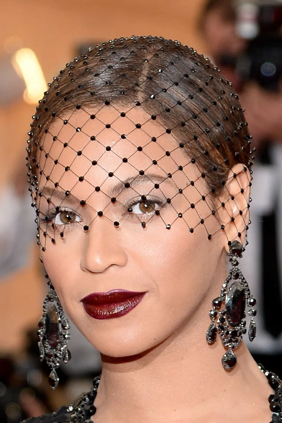Close-up portrait of a woman wearing a black beaded net veil and large chandelier earrings, with bold burgundy lips.