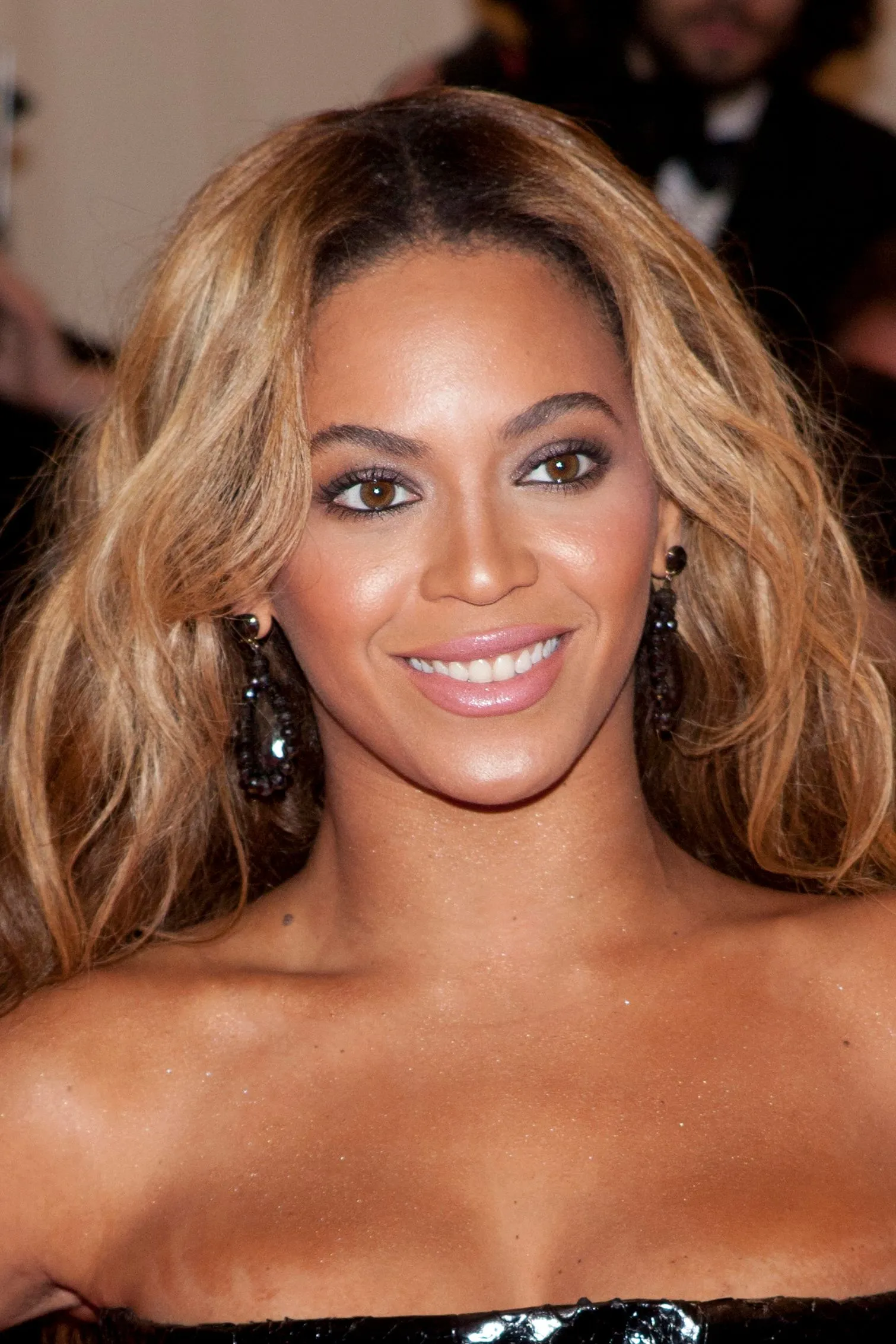 Smiling woman with wavy blonde hair and dark earrings at a formal event (close‑up)