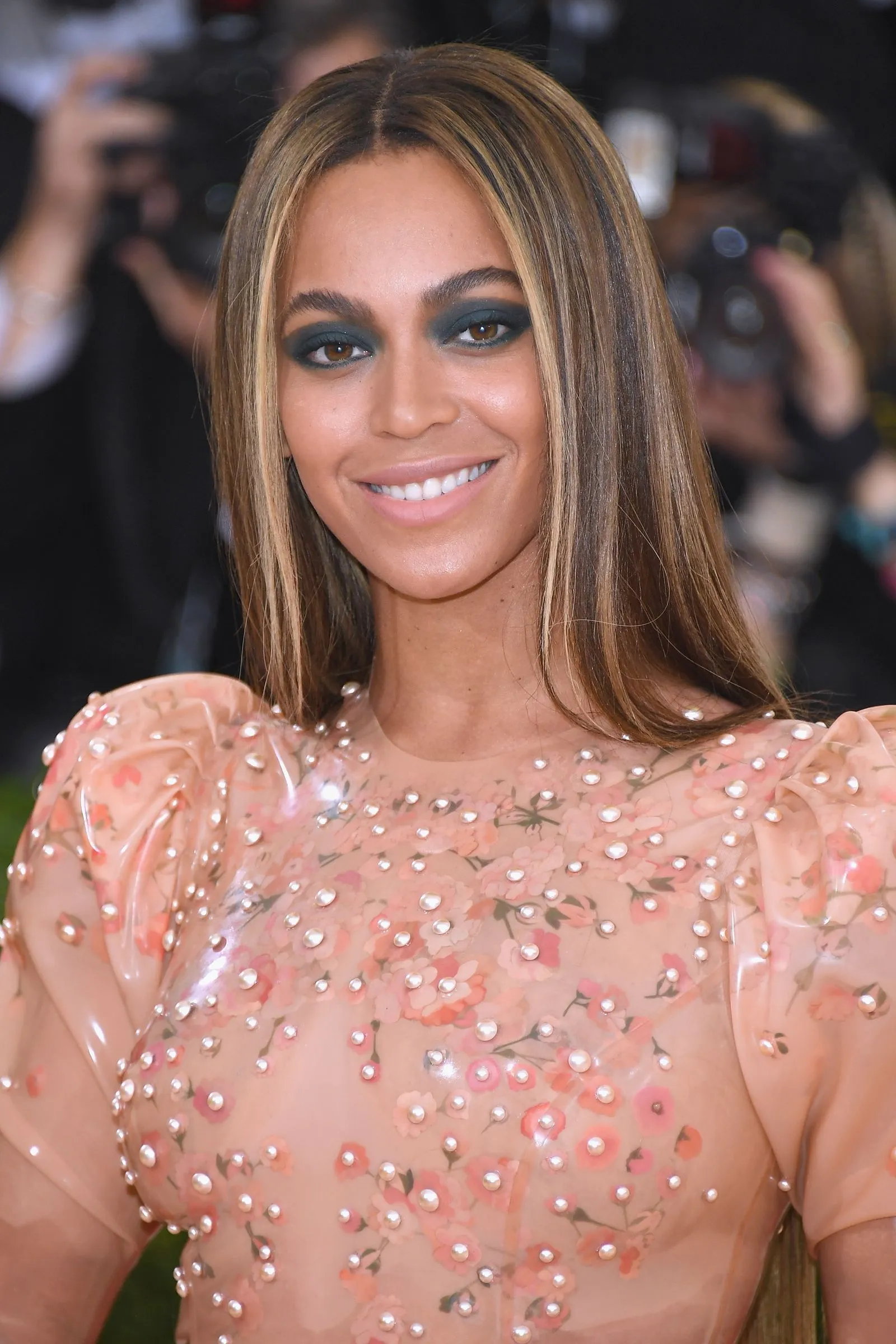 Portrait of a smiling woman with olive skin and teal eye makeup, wearing a sheer pink floral dress with pearl embellishments.