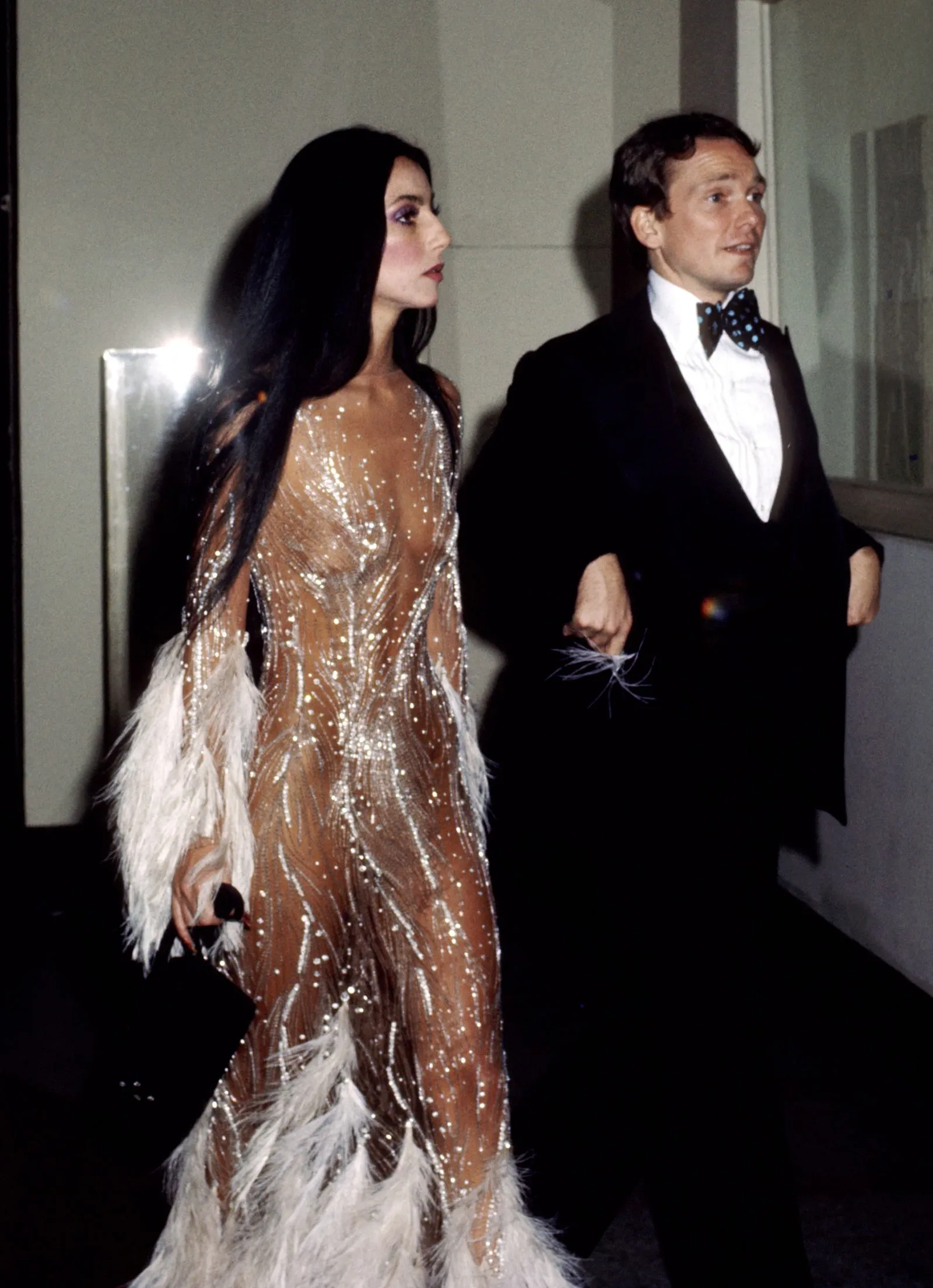 A man in a tuxedo and a woman in a shimmering beige gown walk side by side, entering a room.