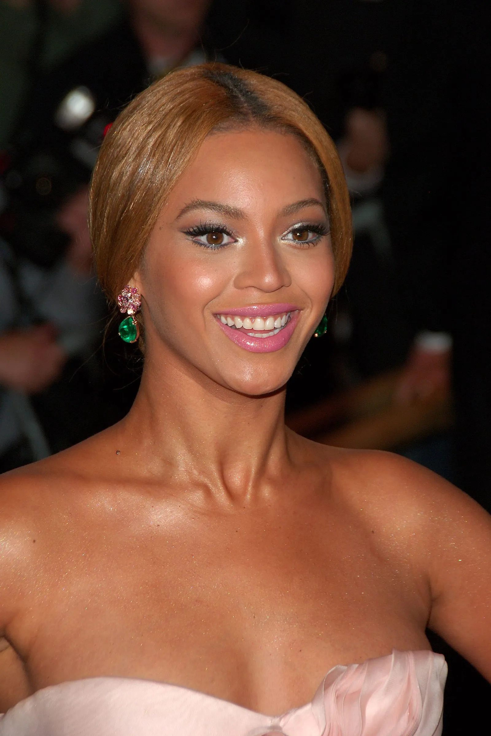Smiling woman with golden-blonde hair, pink lipstick, and green gem earrings.