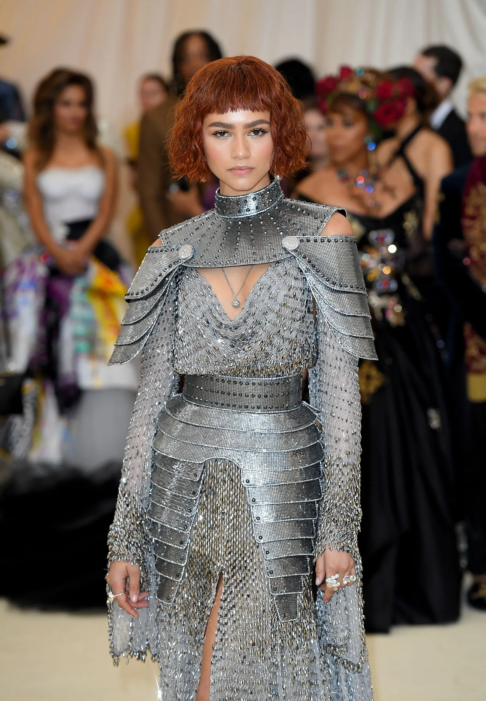 Model wearing a futuristic silver armor-inspired dress with a high neck at a fashion show, crowd in the background.
