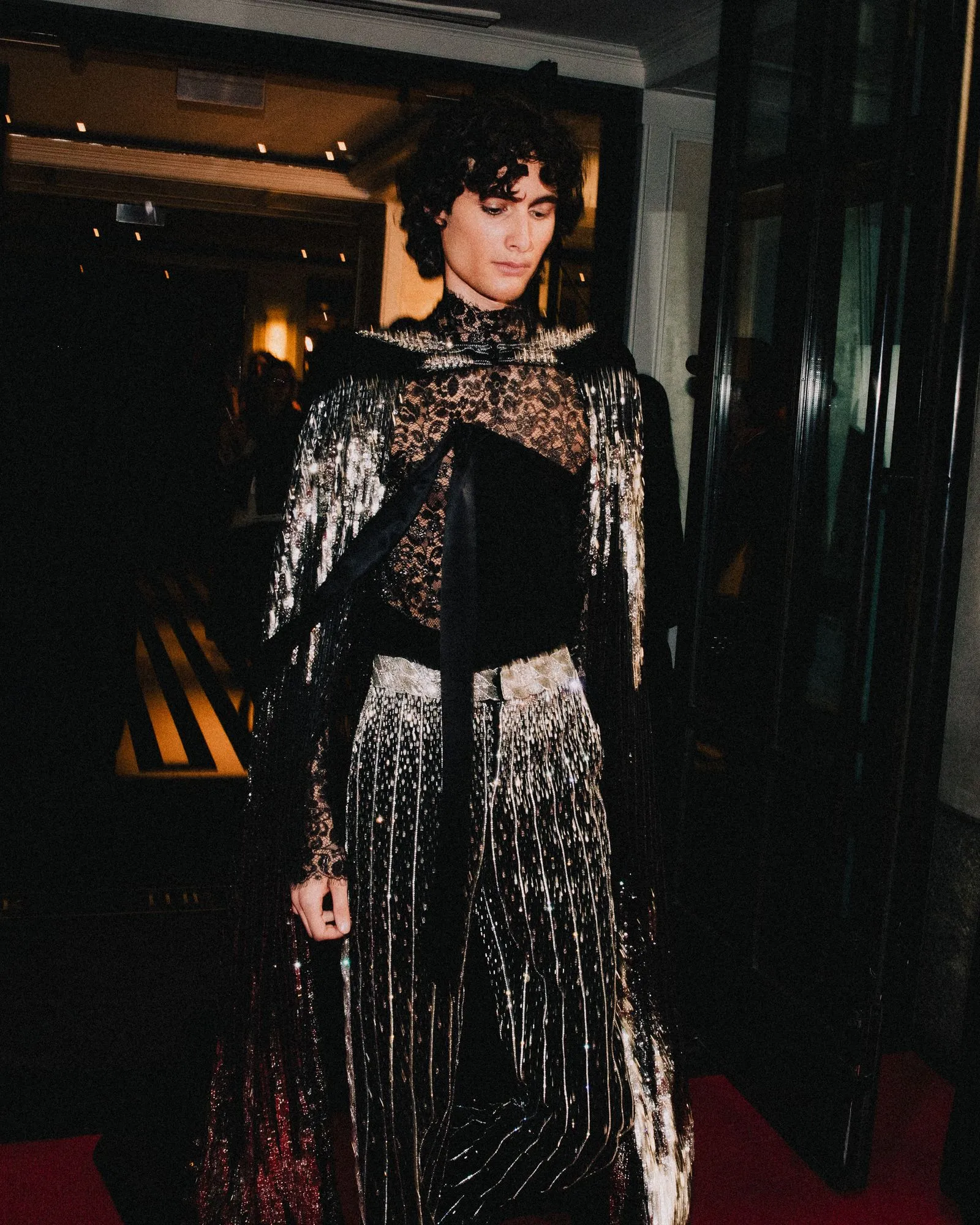 Person in a black lace gown with silver fringe at a red-carpet event, indoors, looking down slightly.