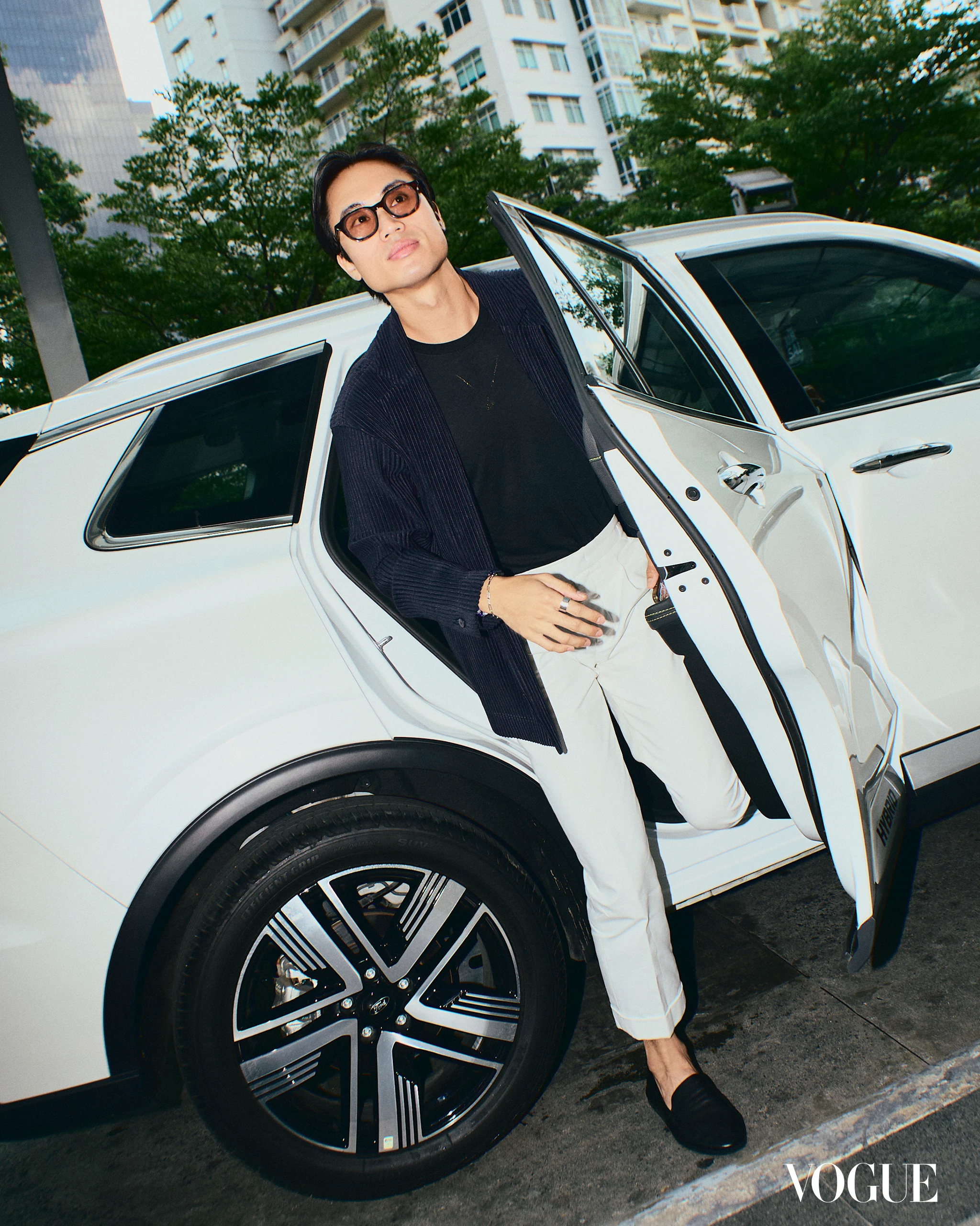Model steps out of a white luxury SUV wearing a dark jacket, black top, white pants, and sunglasses against an urban backdrop; Vogue watermark visible.