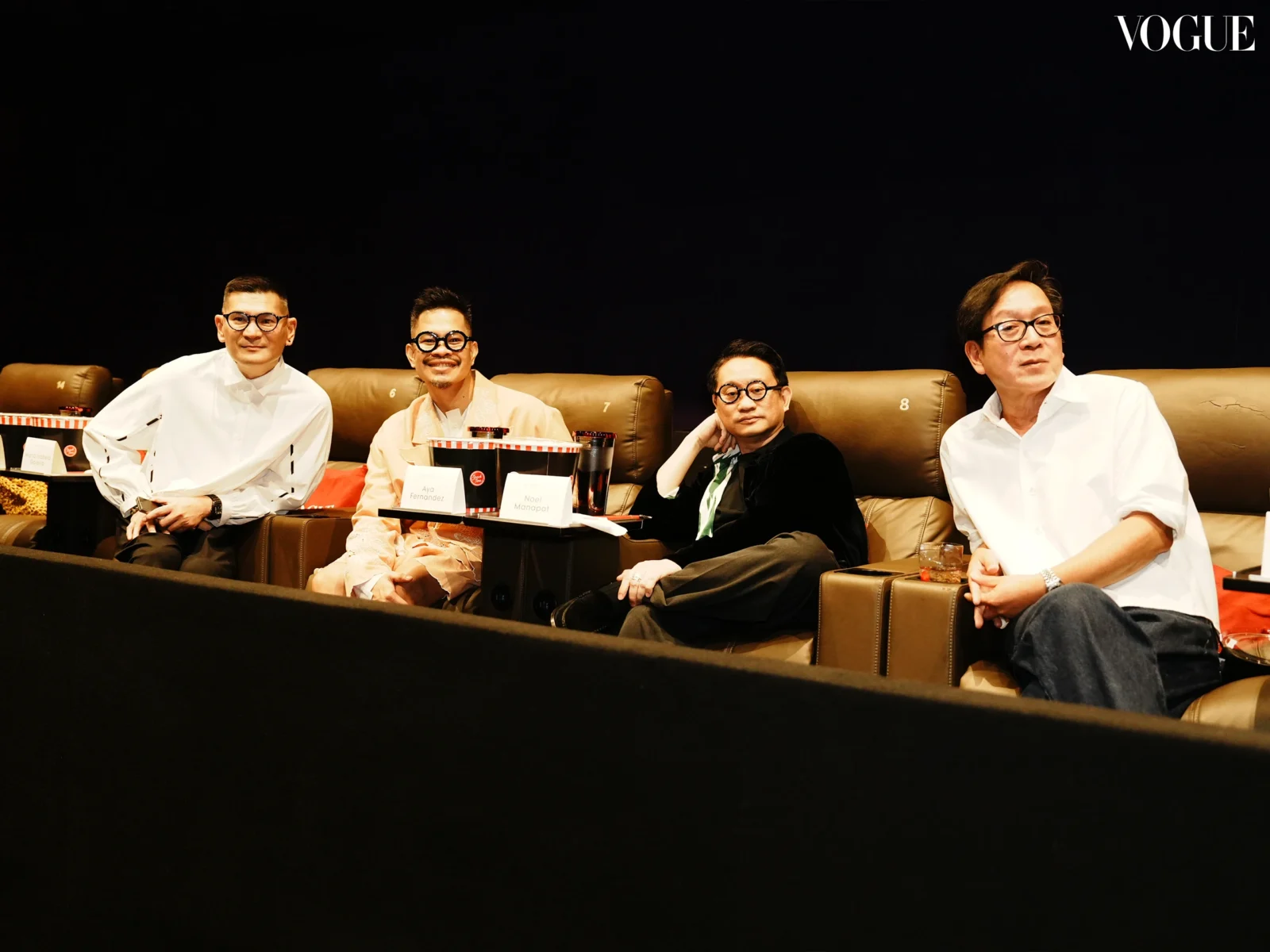 Four men sit on a brown leather couch on a stage, wearing white shirts and glasses, with nameplates in front, at a Vogue event.