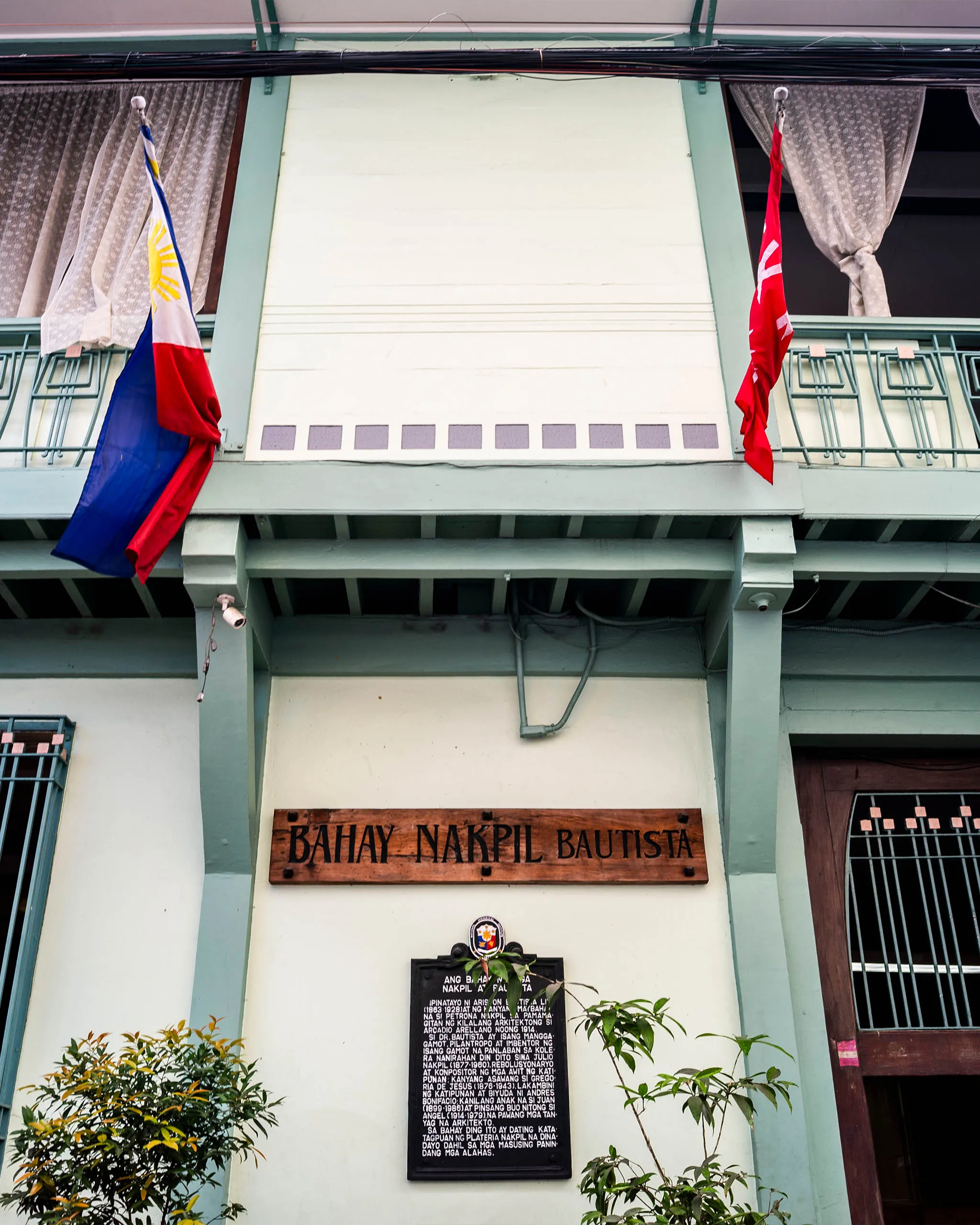 Facade of a building with two flags (Philippines flag on the left, red flag on the right), green railings, and a wooden plaque reading 'BAHAY NAKPIL BAUTISTA' with surrounding plants.