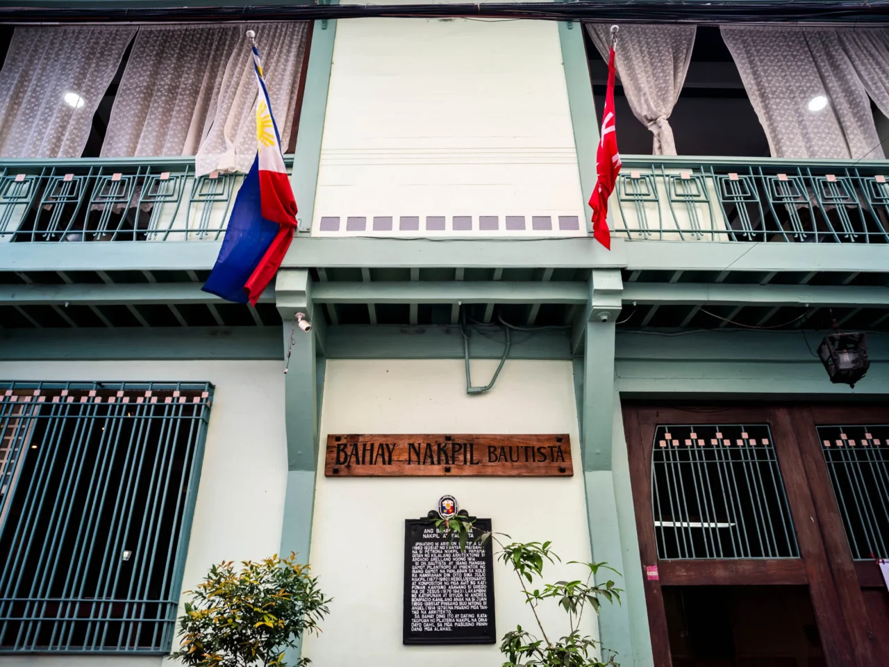 Facade of a historic building with turquoise railings, flags, and a wooden plaque reading 'BAHAY NAKPIL BAUTISTA' above the entrance.