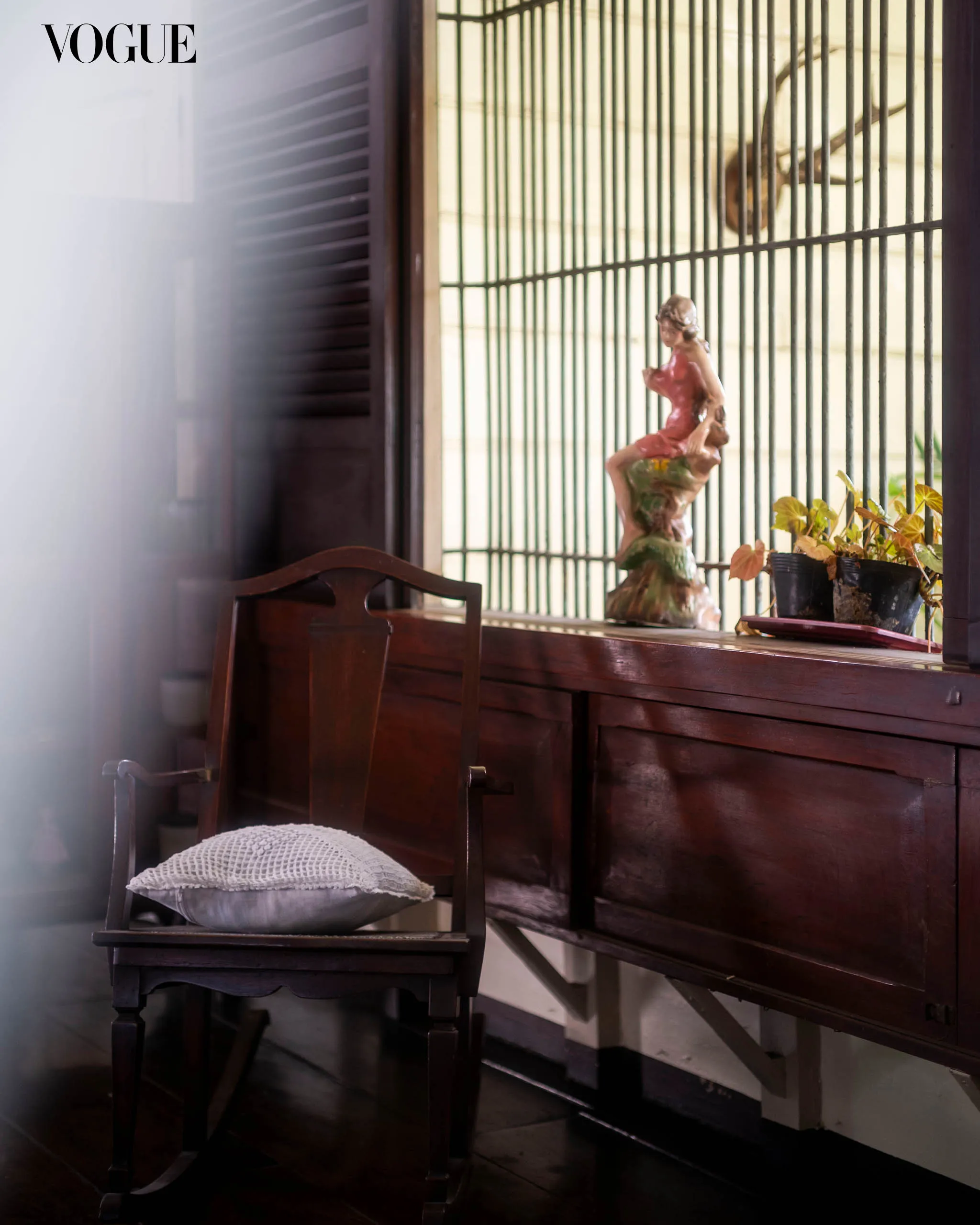 Vogue logo in the top-left corner with a vintage wooden piano and bench, a decorative statue, and potted plants behind iron window bars in a sunlit room.