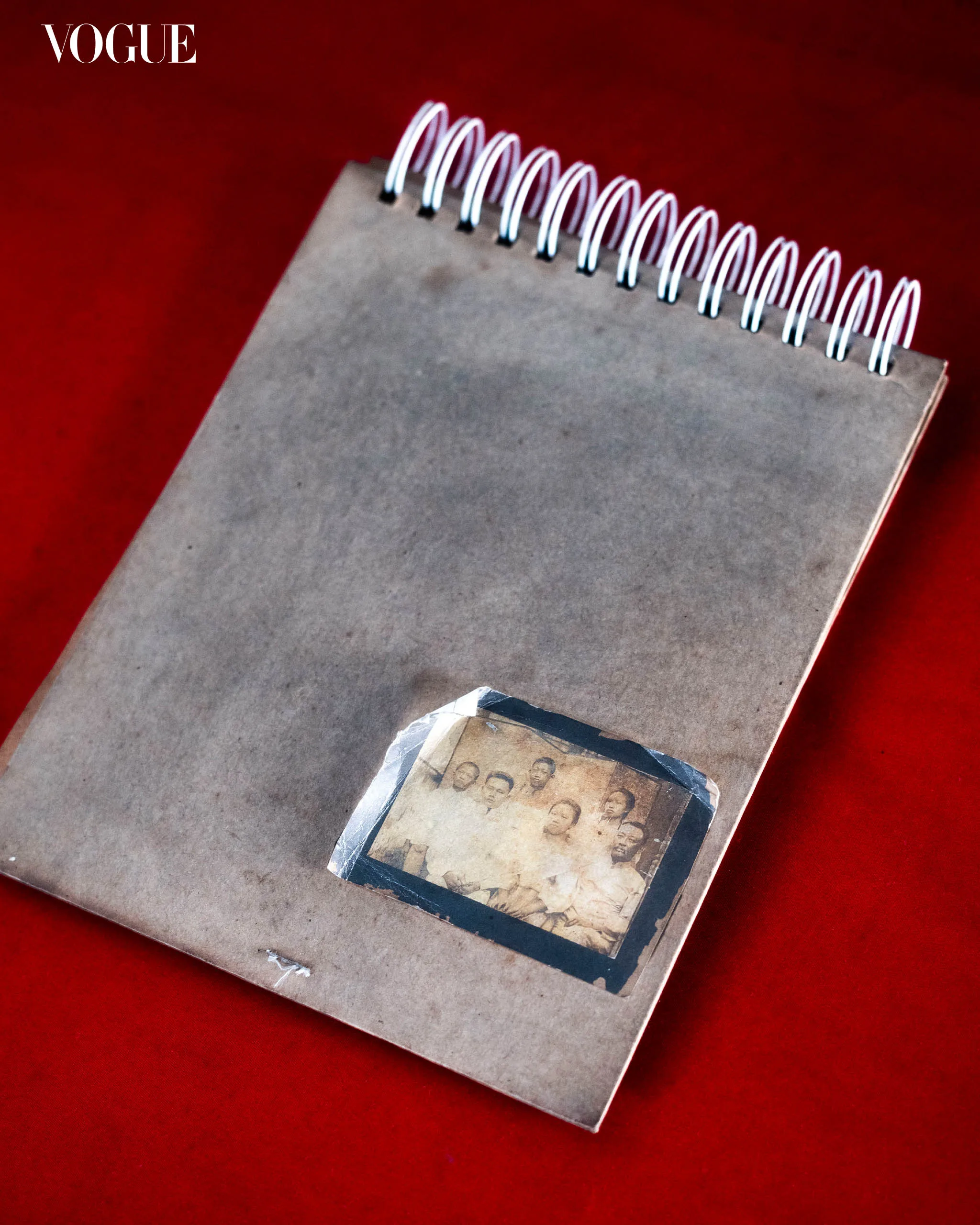 Notebook on a red surface with a white spiral binding; a small vintage photo collage is taped to the cover and the Vogue logo is in the corner.