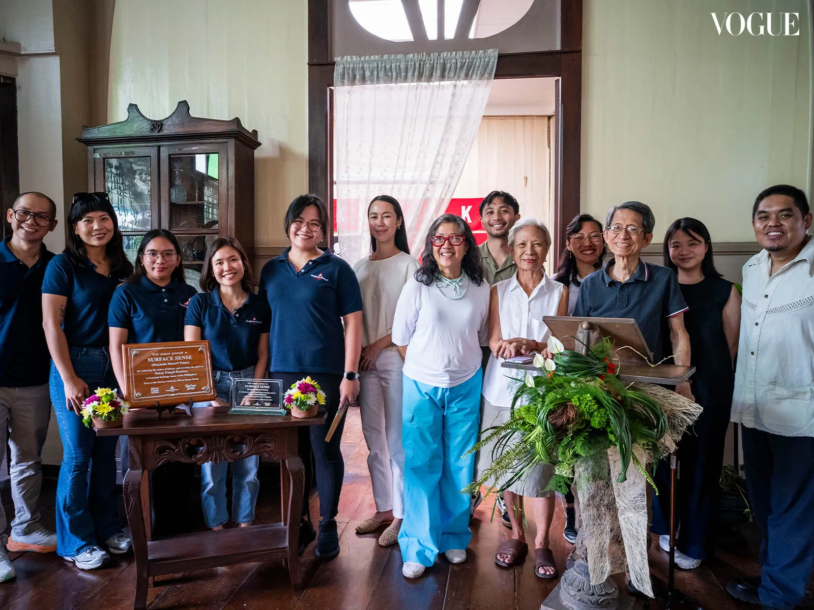Group of about a dozen people posing for a photo indoors, some holding plaques, with a wooden table and floral centerpiece in front.
