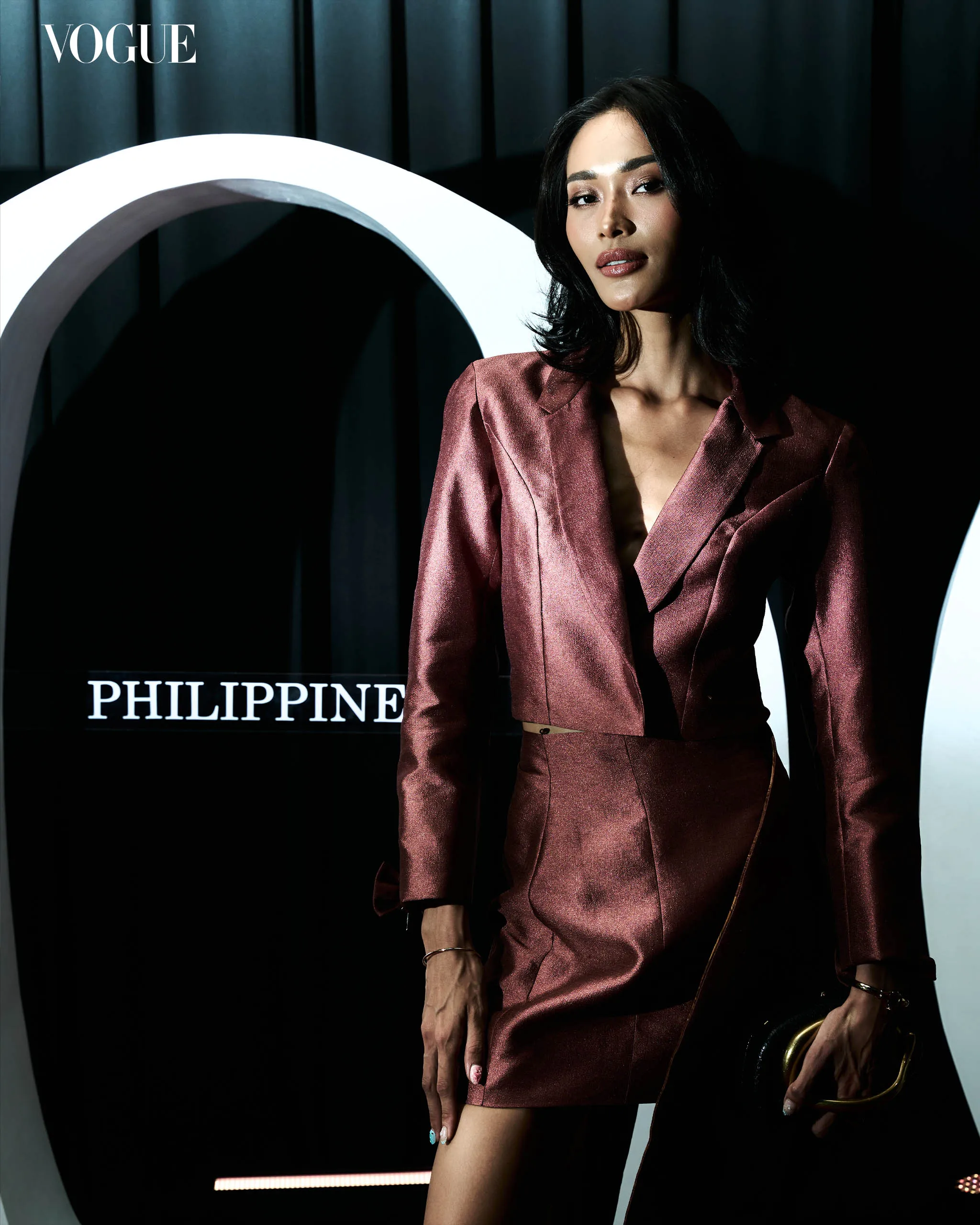 Model in a metallic burgundy blazer and skirt posed beside a large white curved sculpture on a dark backdrop, with Vogue Philippines branding.