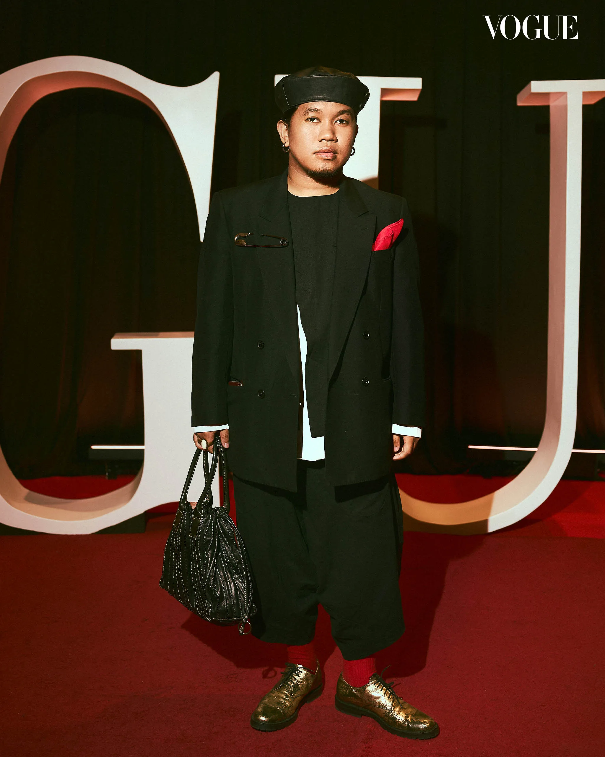 Person wearing a black suit with a red pocket square and beret on a red carpet at a Vogue event, holding a black striped handbag and wearing metallic gold shoes with red socks.