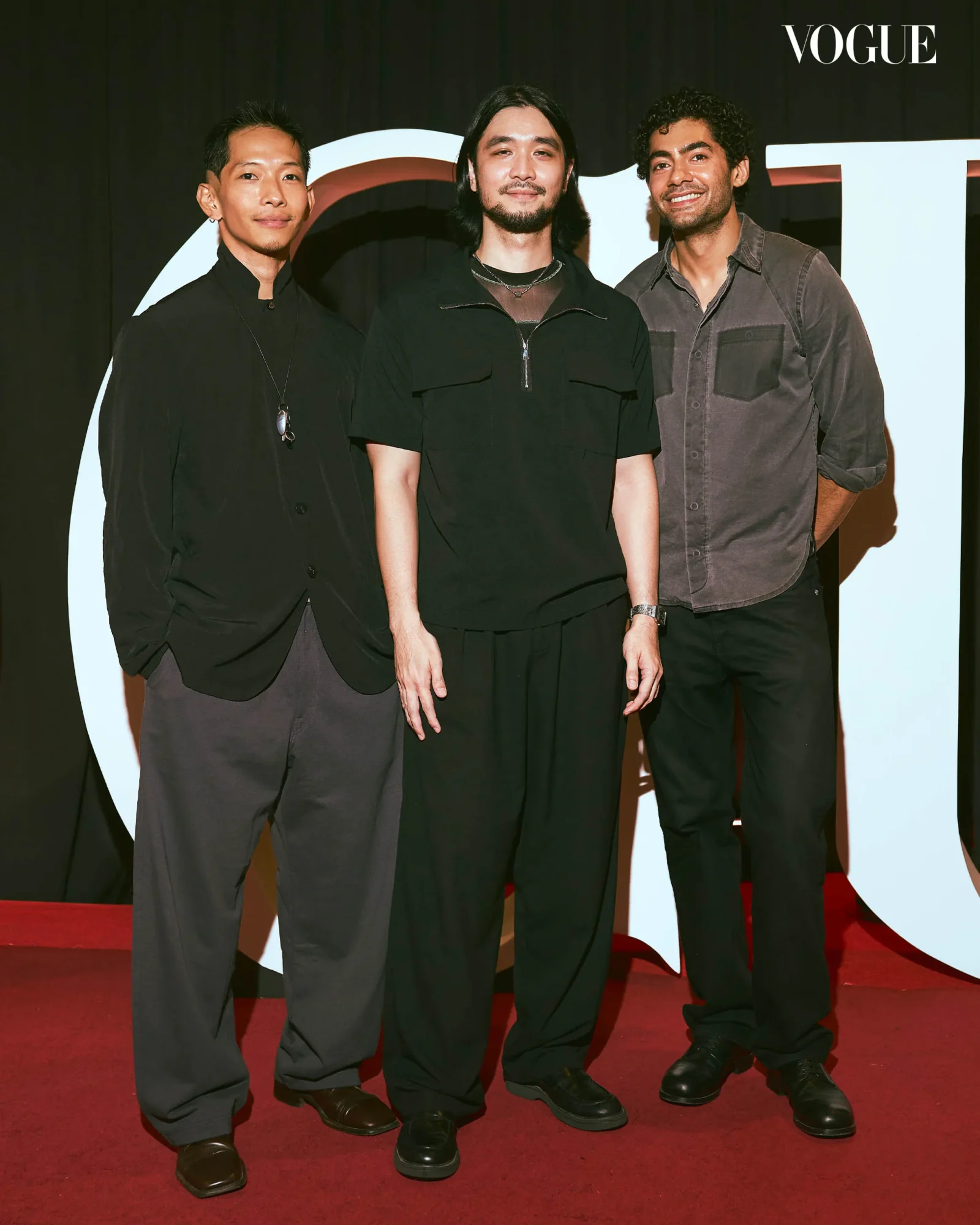 Three men pose on a red carpet at a Vogue event, wearing dark outfits, with a large white backdrop behind them.