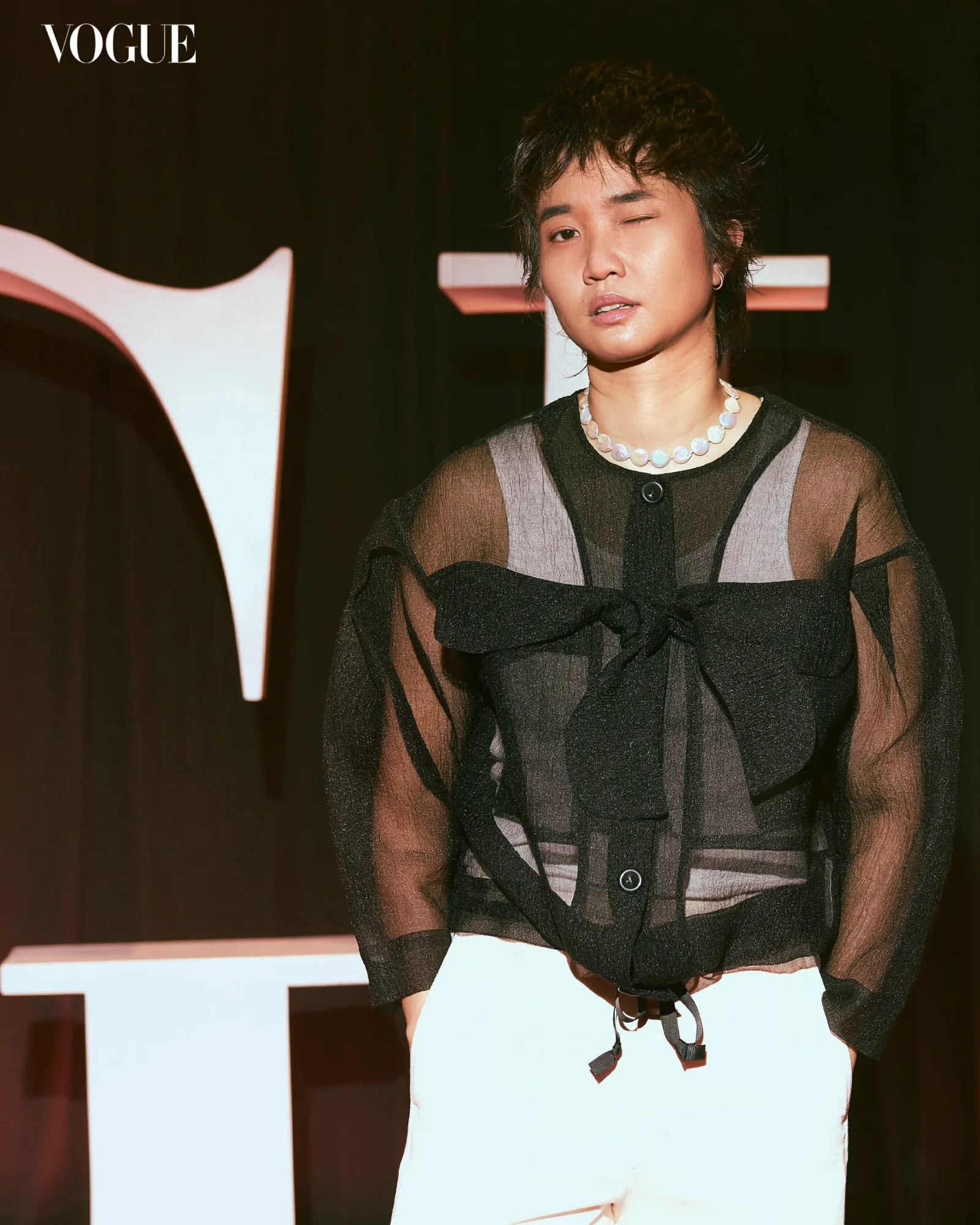 Person in a sheer black blouse with a pearl necklace poses beside a large white sign at a Vogue event; Vogue logo is in the top left corner.