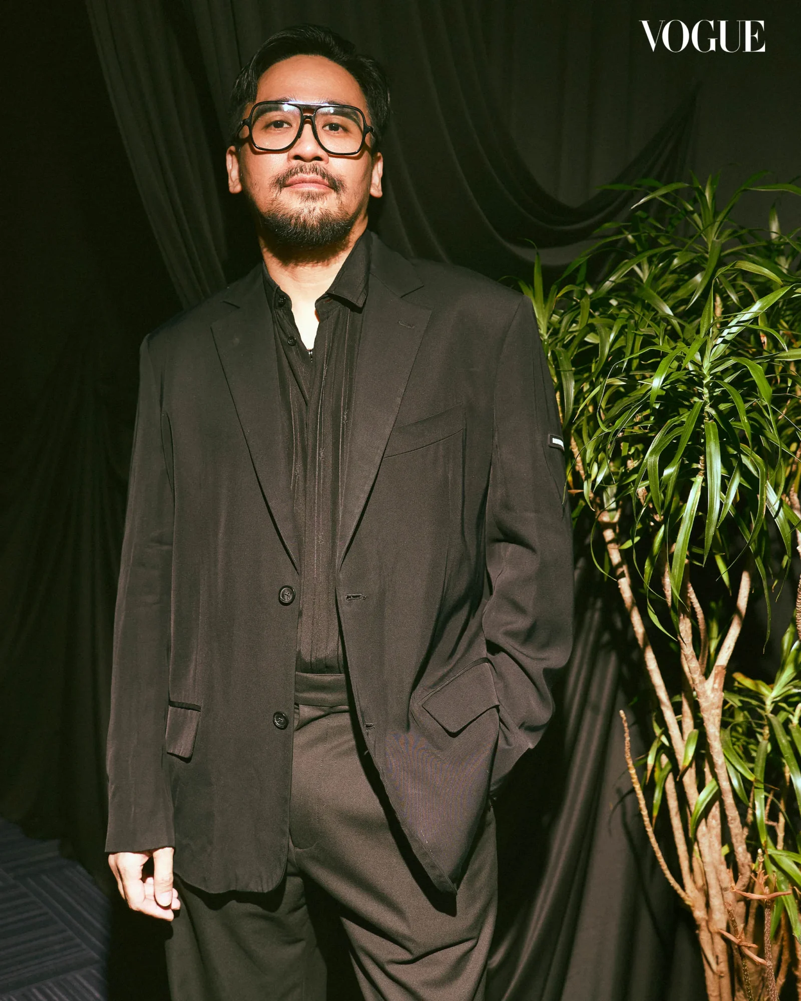 Portrait of a man in a black suit and large glasses posed against a black backdrop with green plants to the right; Vogue logo in the corner.