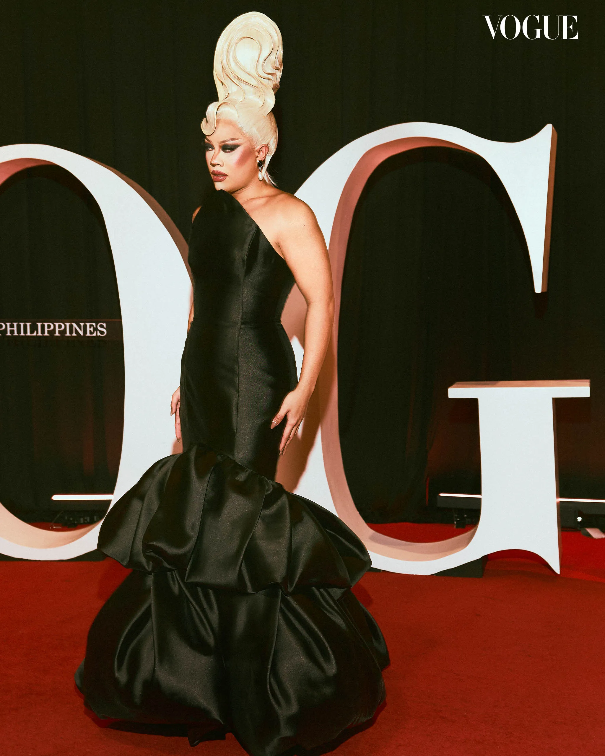 Person in a sleek black gown with a dramatic tall blonde updo poses on a red carpet, Vogue backdrop behind.