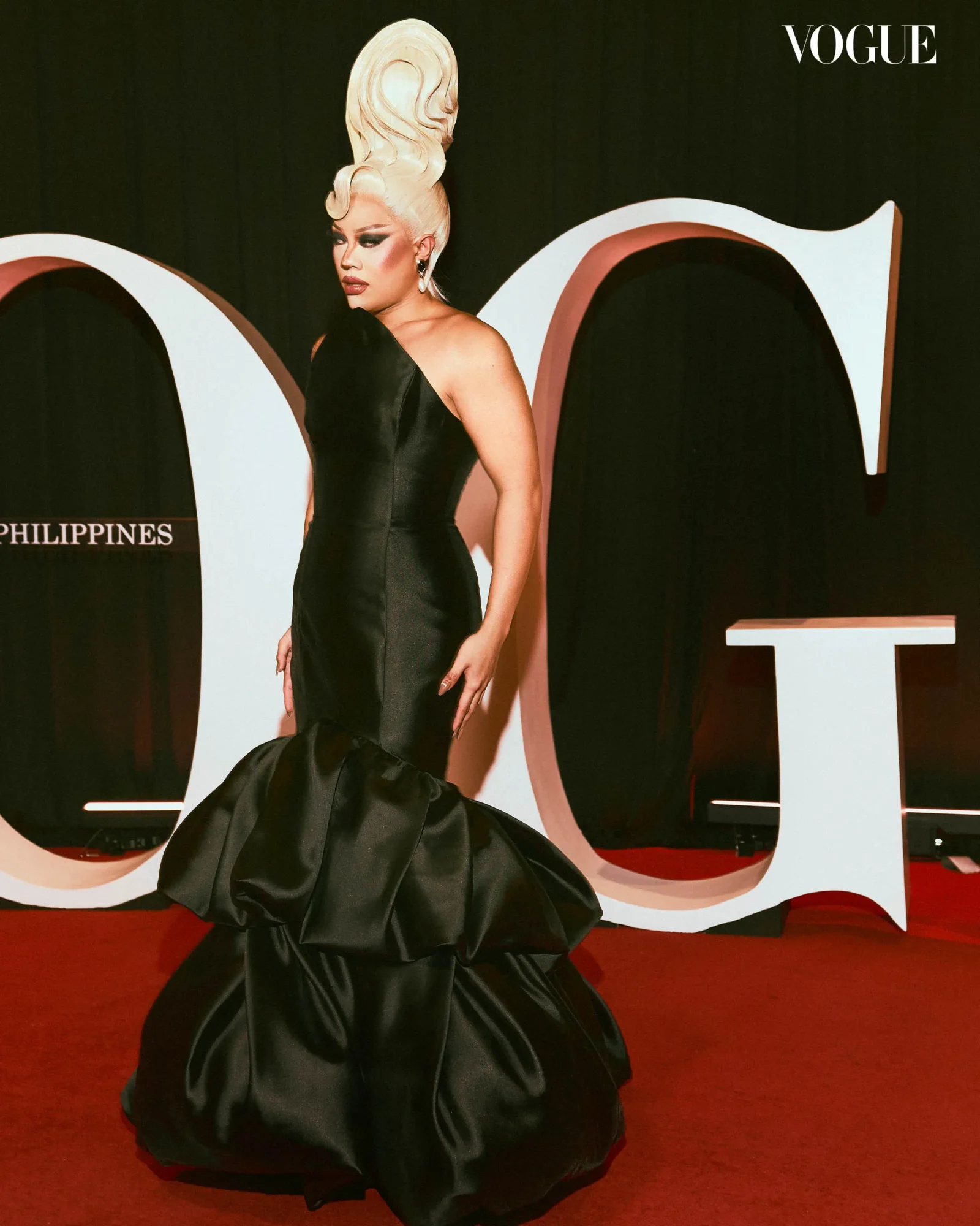 Drag queen in a sleek black evening gown on a red carpet with bold makeup and an extreme updo, Vogue backdrop behind.
