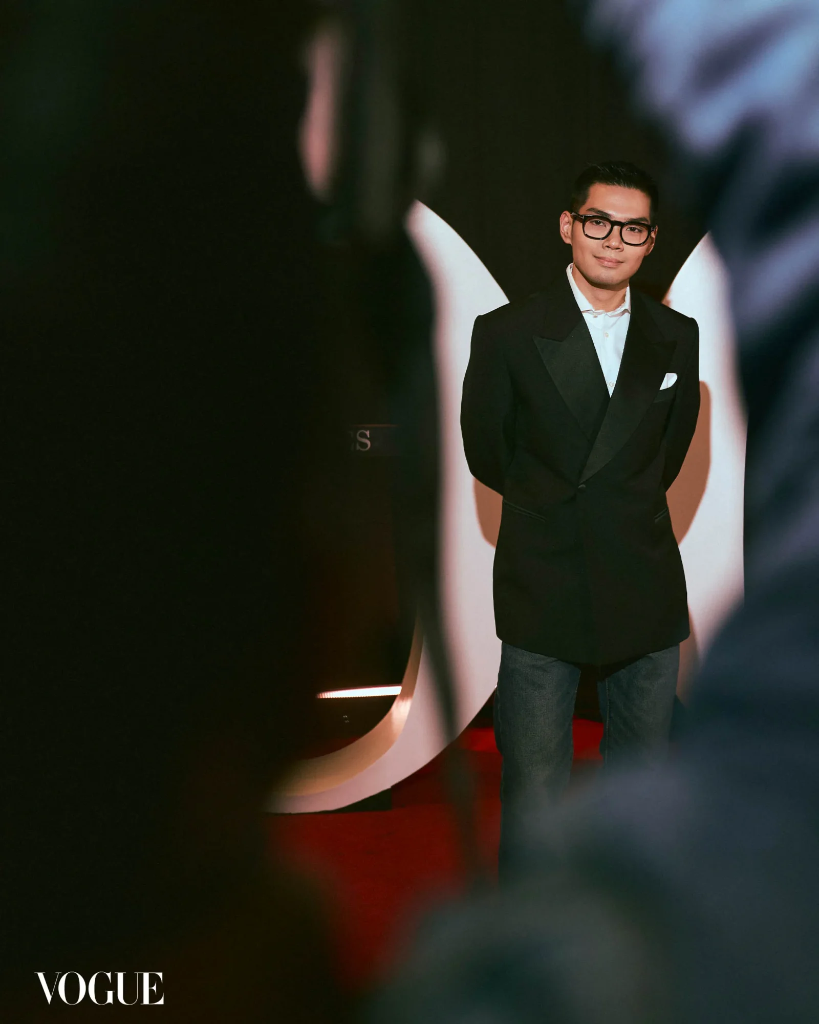 Man in a black tuxedo and glasses posing on a red carpet, Vogue watermark bottom left, soft foreground blur in the foreground.